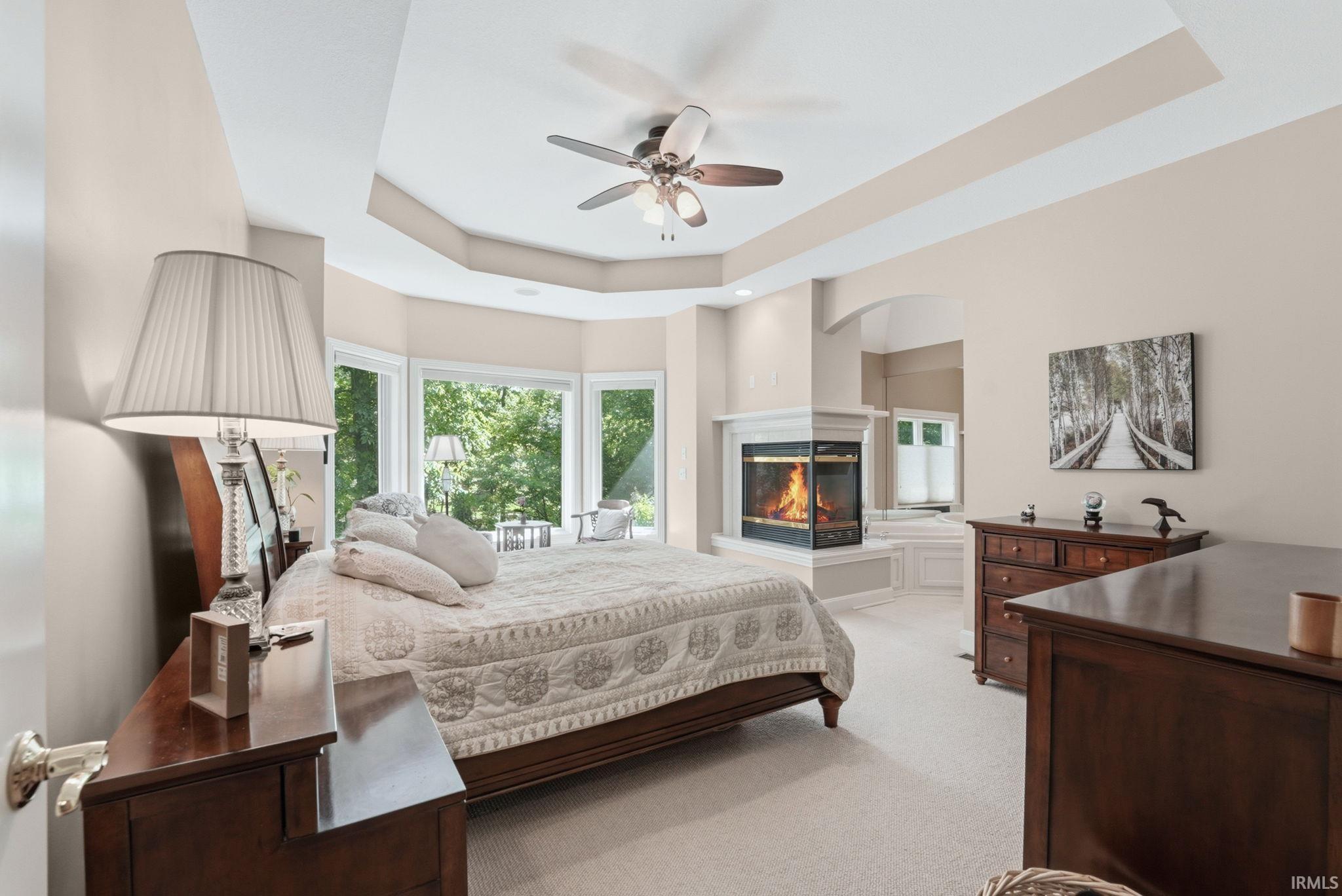 Spacious room featuring a corner fireplace with white surround, recessed tray ceiling, ceiling fan, and bay windows
