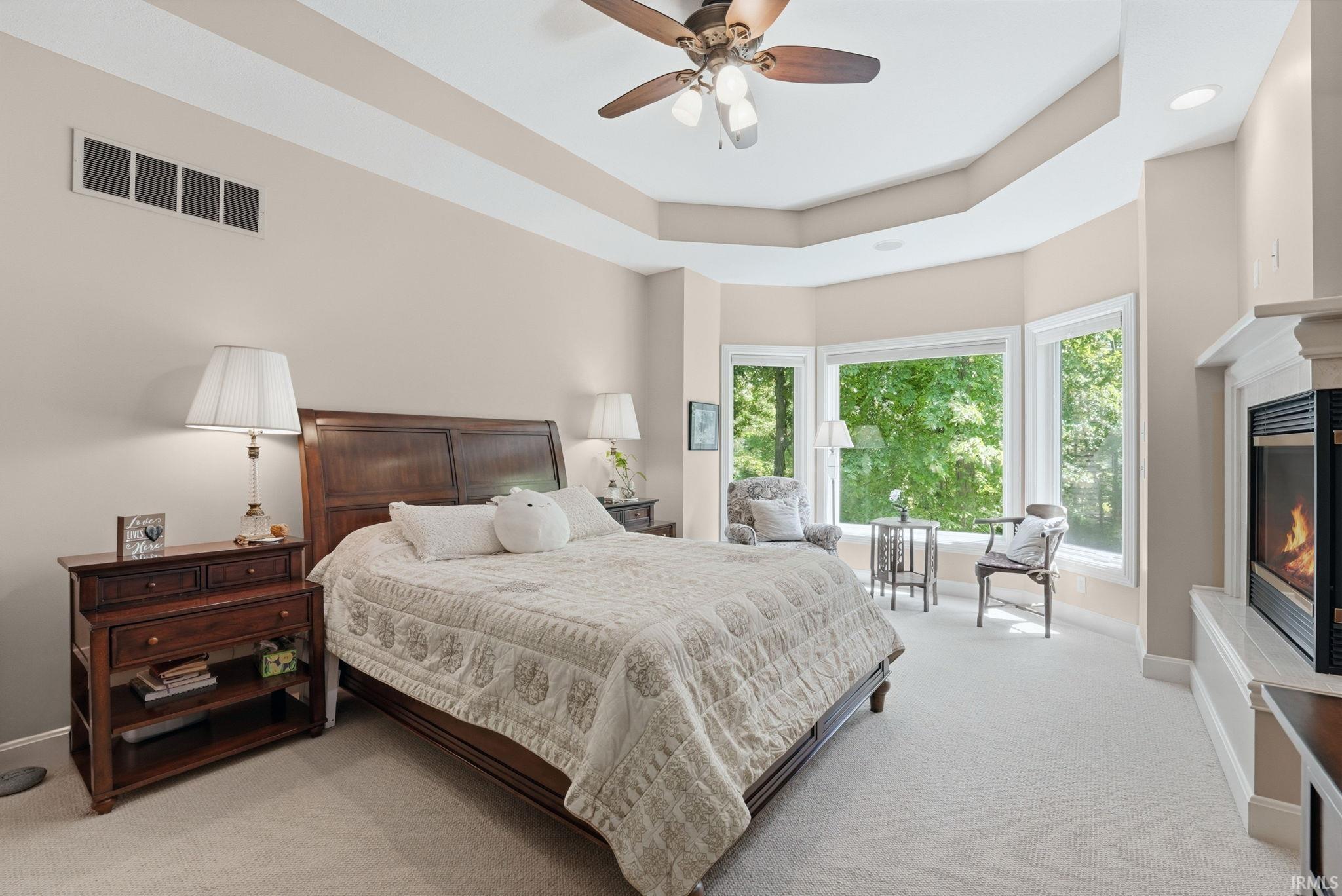 Spacious bedroom featuring a tray ceiling, ceiling fan, carpeting, a bay window with multiple panes, and a built-in fireplace
