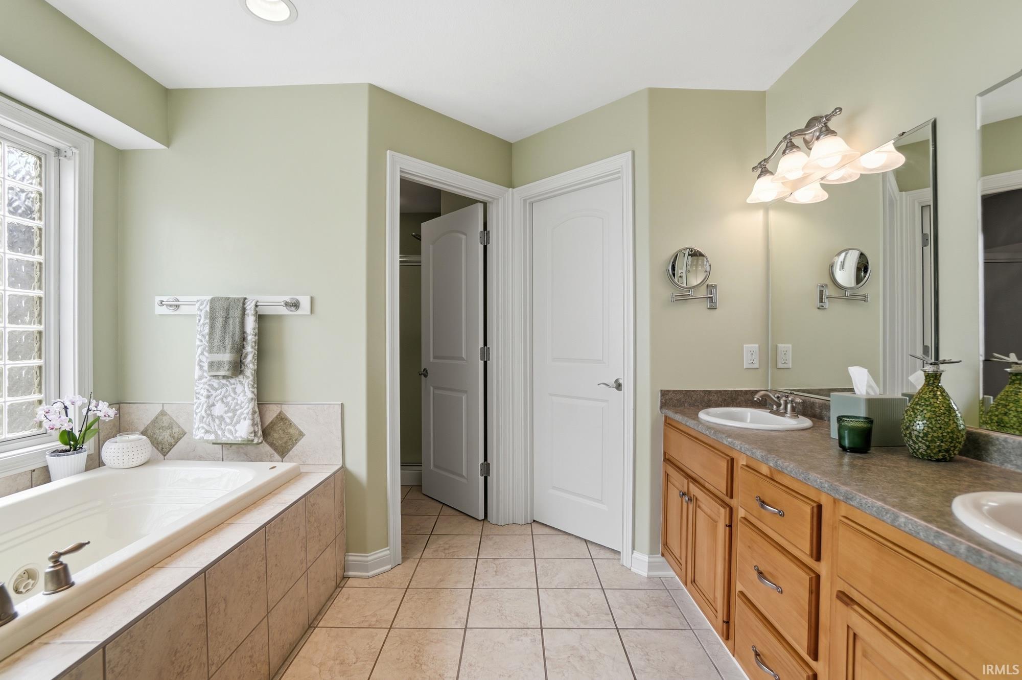 Bathroom featuring a built-in soaking tub with tile surround, a double vanity with light wood cabinetry and integrated sinks, and tiled flooring