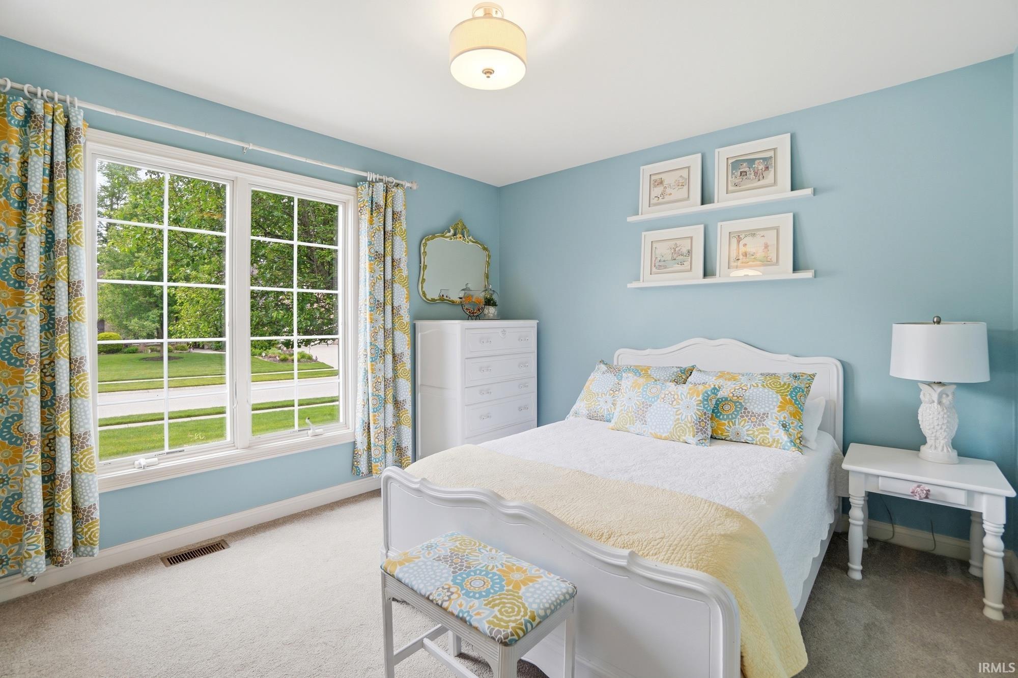 Light blue bedroom featuring a large picture window with grid patterns, ceiling light fixture, wall shelving, and neutral carpeting