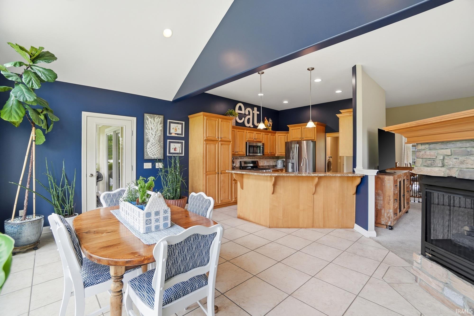 Open-concept interior featuring light-toned tile flooring, a kitchen with natural wood cabinetry and stainless steel appliances, and a built-in fireplace with a stone surround