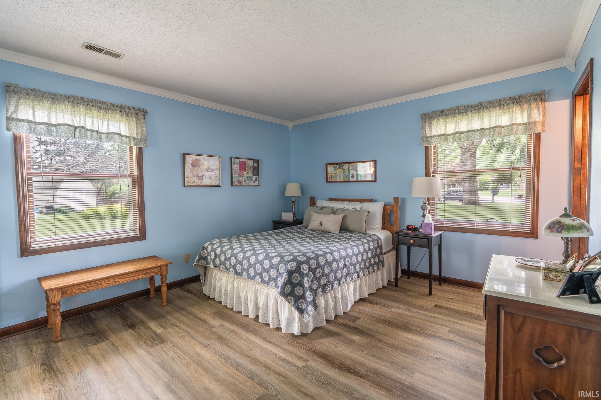 Spacious room featuring wood-finish flooring, two large windows with blinds, crown molding, and light blue walls