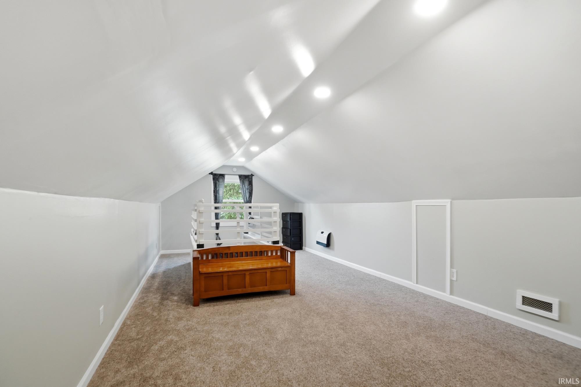 Spacious carpeted room featuring a vaulted ceiling with recessed lighting, light gray walls, and a window with dark drapes