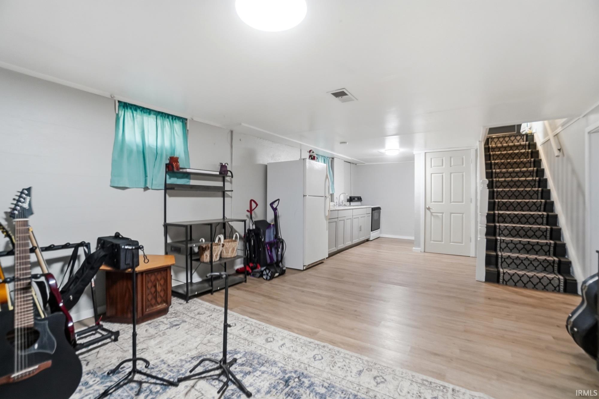 Spacious lower-level area featuring light wood-finish flooring, a kitchen area with white cabinetry and a white refrigerator, a staircase with patterned carpeting, and white walls