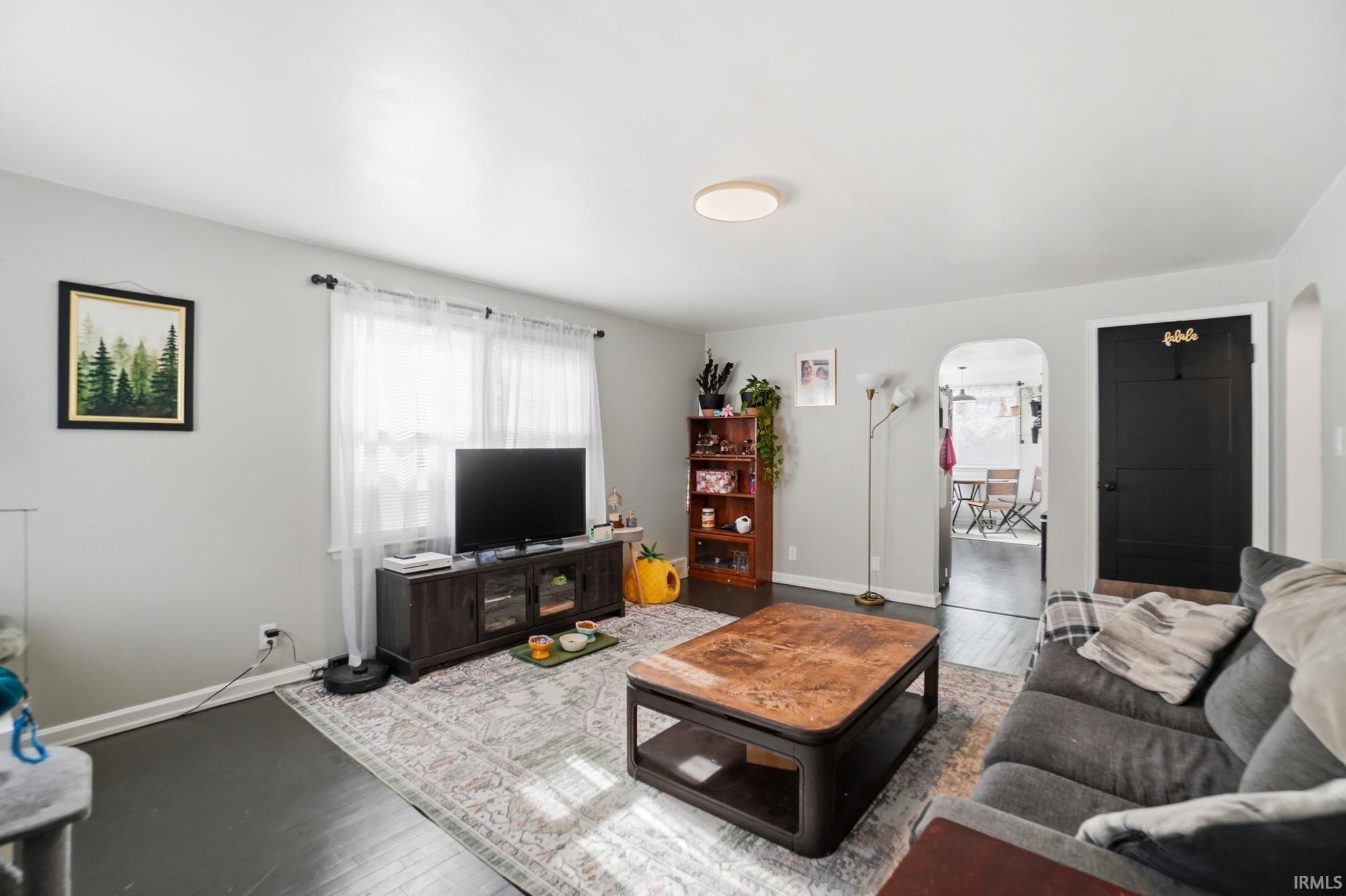Living area featuring dark wood-finish flooring, light gray walls, and a large window with white sheer curtains