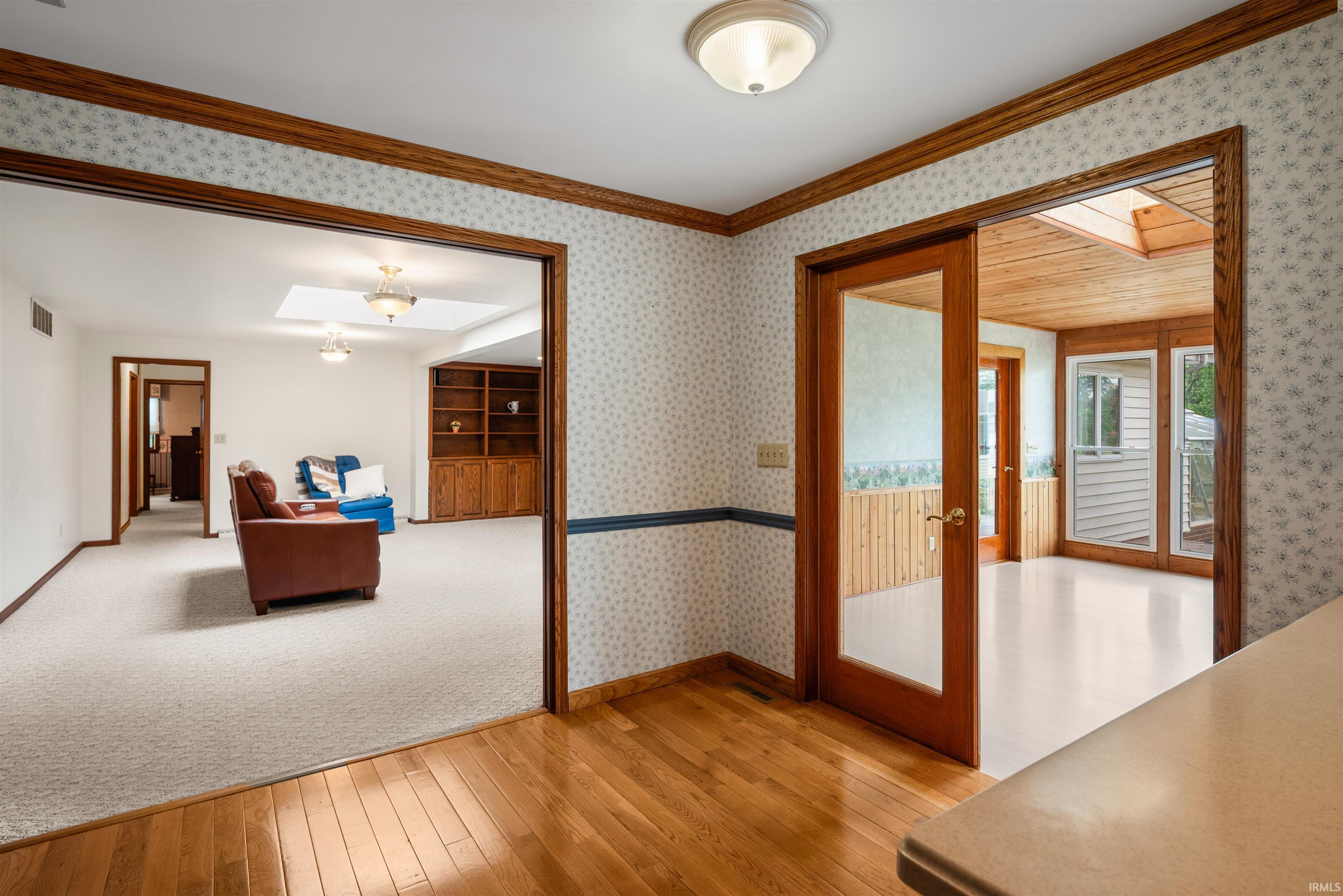 Hardwood flooring present in the foreground, with an entryway featuring a glass-panel door opening into a room with wood-paneled walls and a wood-slat ceiling with a skylight