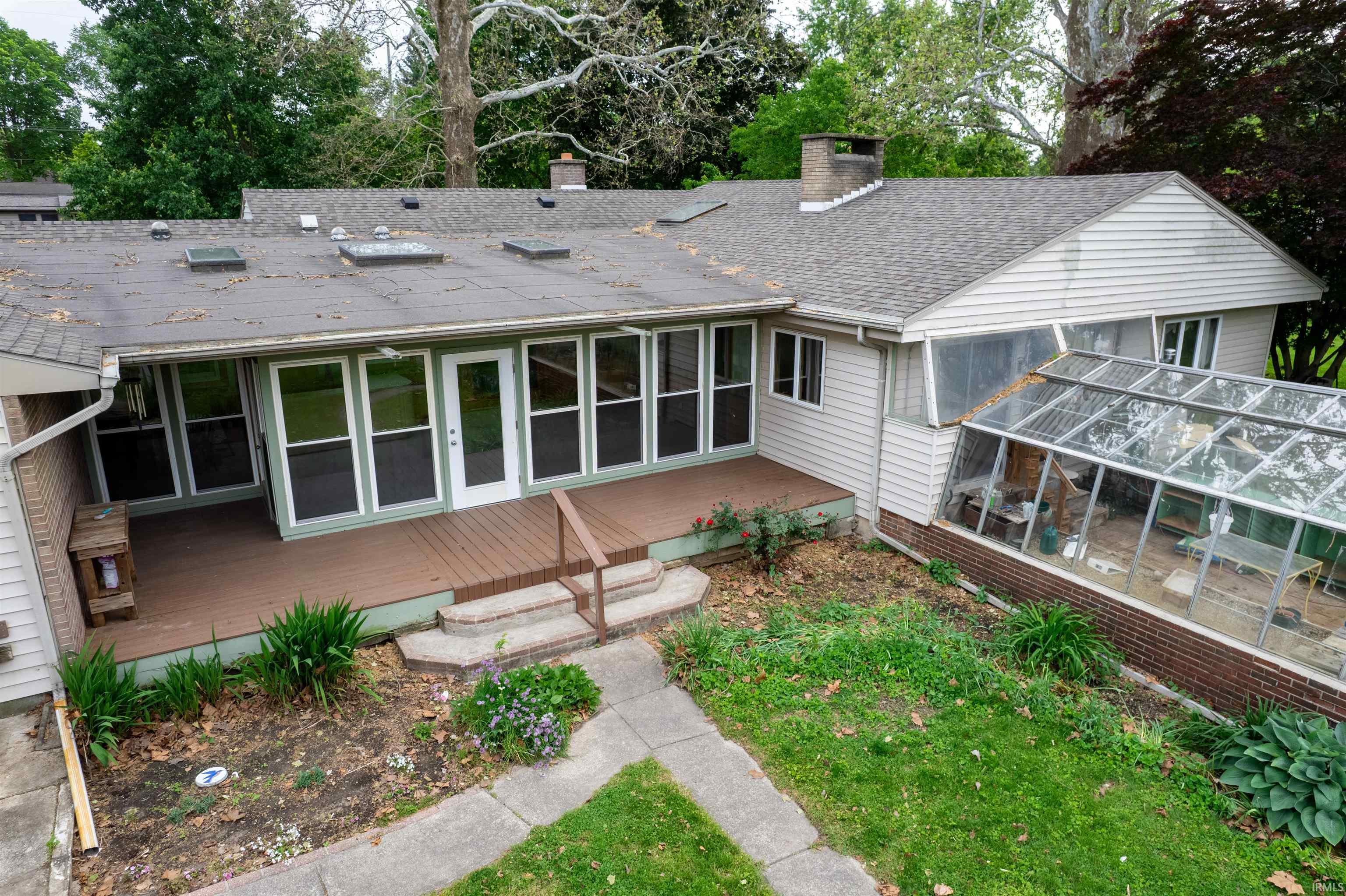 Rear exterior featuring a sunroom with white-framed windows, a wood-finish deck with railing, and an attached greenhouse with a brick base