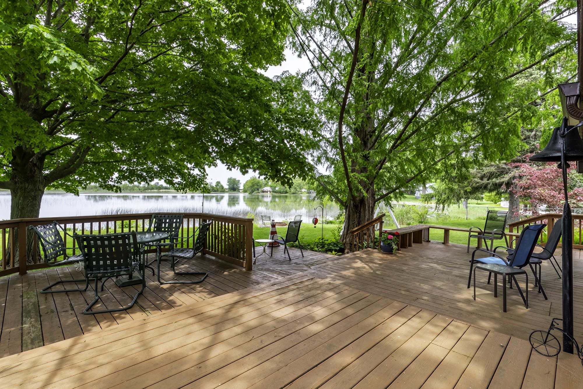 Expansive outdoor wood-finish deck with wood railing, mature trees providing natural shade, and a visible water body