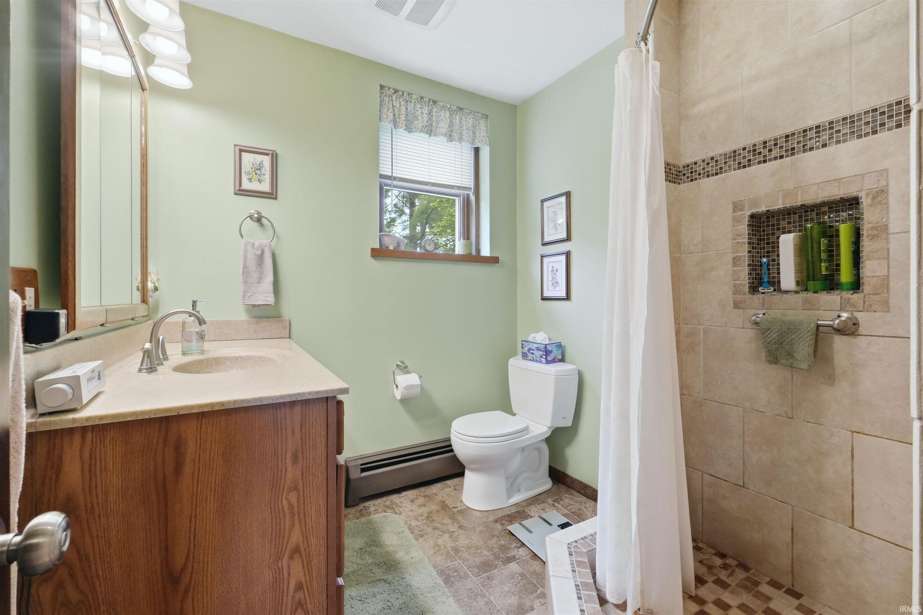 Bathroom featuring a wood-finish vanity with a light-toned countertop, a tiled shower with an integrated niche and mosaic accent, and a window with blinds