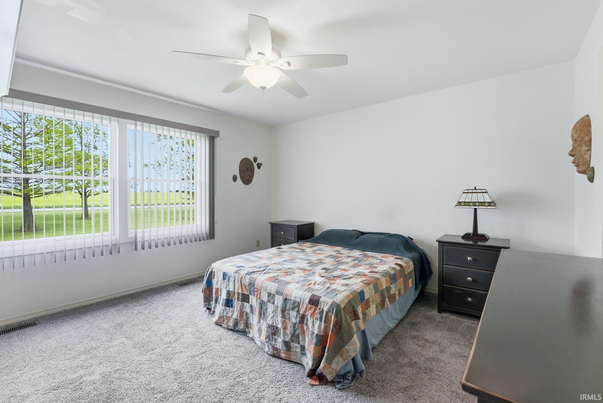 Bright interior space featuring a large window with vertical blinds, a ceiling fan with integrated lighting, and neutral-toned wall-to-wall carpeting