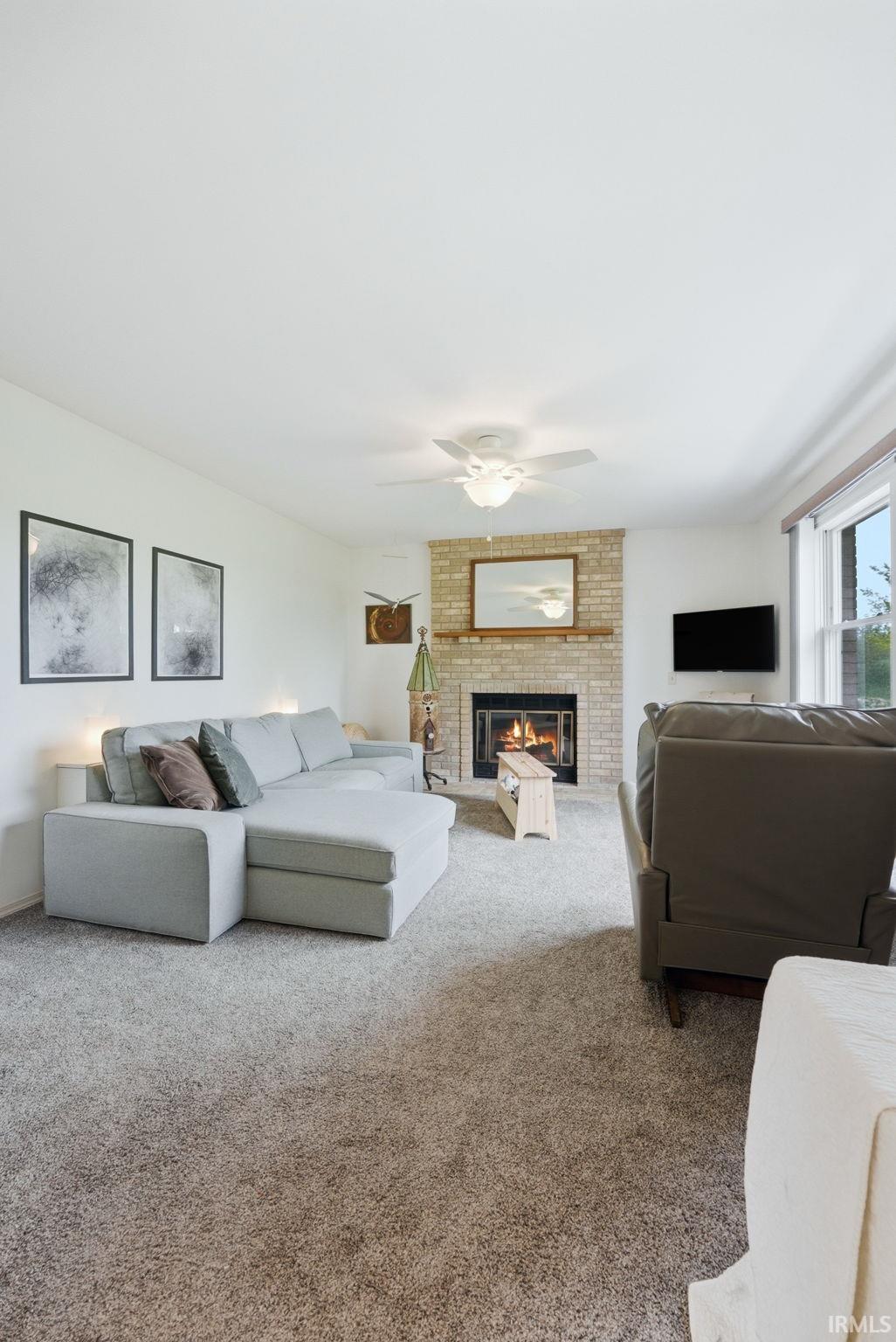 Spacious room featuring a brick fireplace with a wood mantle, soft neutral carpeting, a ceiling fan, and natural light from a window