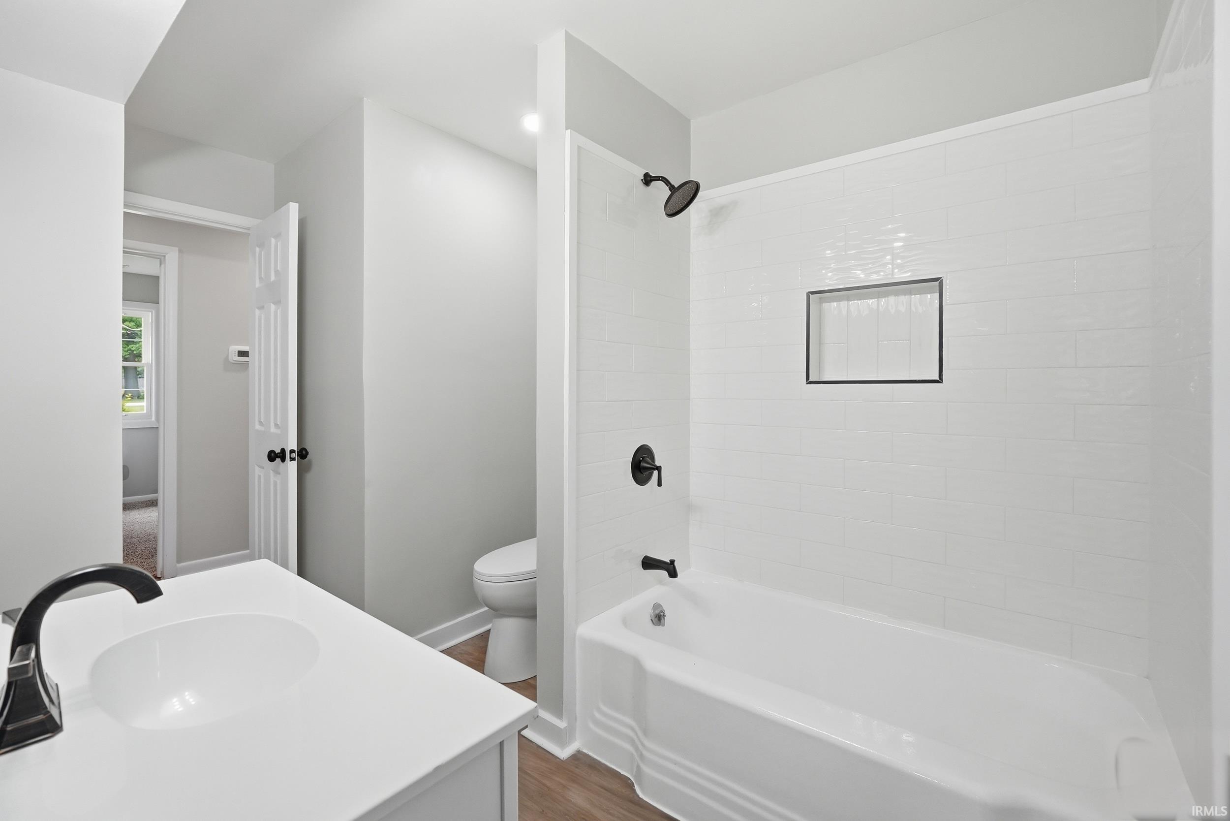 Bathroom featuring a white vanity with an integrated sink and black faucet, a white subway-tiled shower/tub combination with a recessed niche and black fixtures, and wood-finish flooring