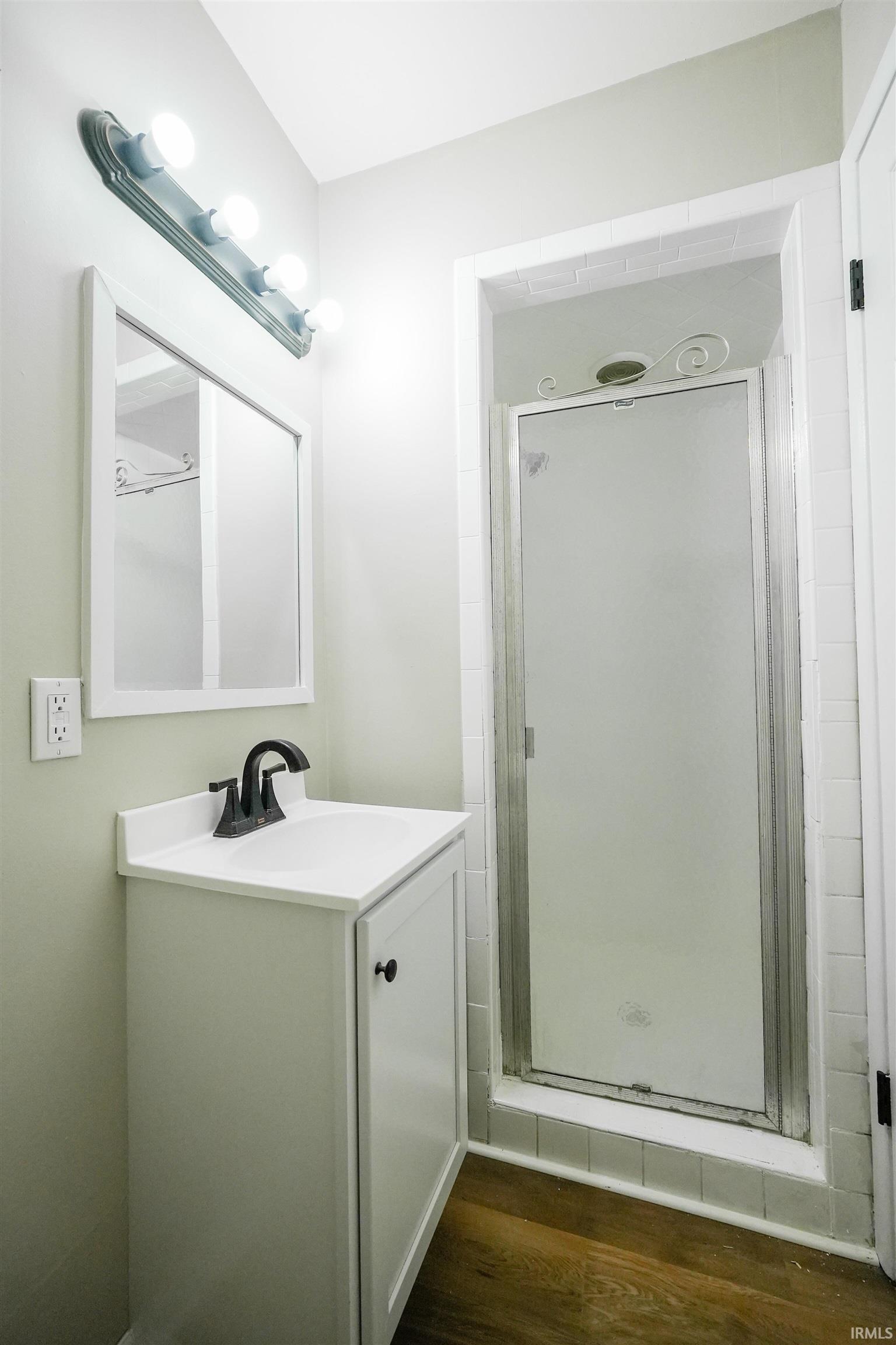 Compact bathroom featuring a shower stall with a frosted glass door, a white vanity with a dark faucet, a mirrored medicine cabinet, and wood-finish flooring