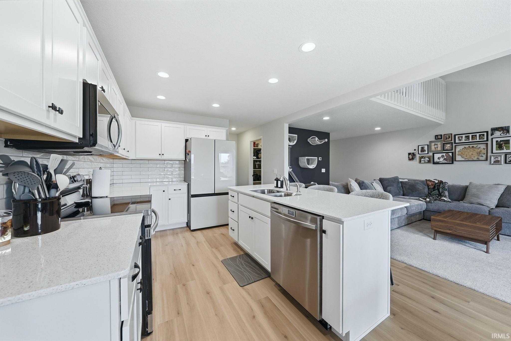 Open-concept kitchen featuring white cabinetry, subway tile backsplash, light-toned wood-finish flooring, a kitchen island with integrated sink, and stainless steel appliances