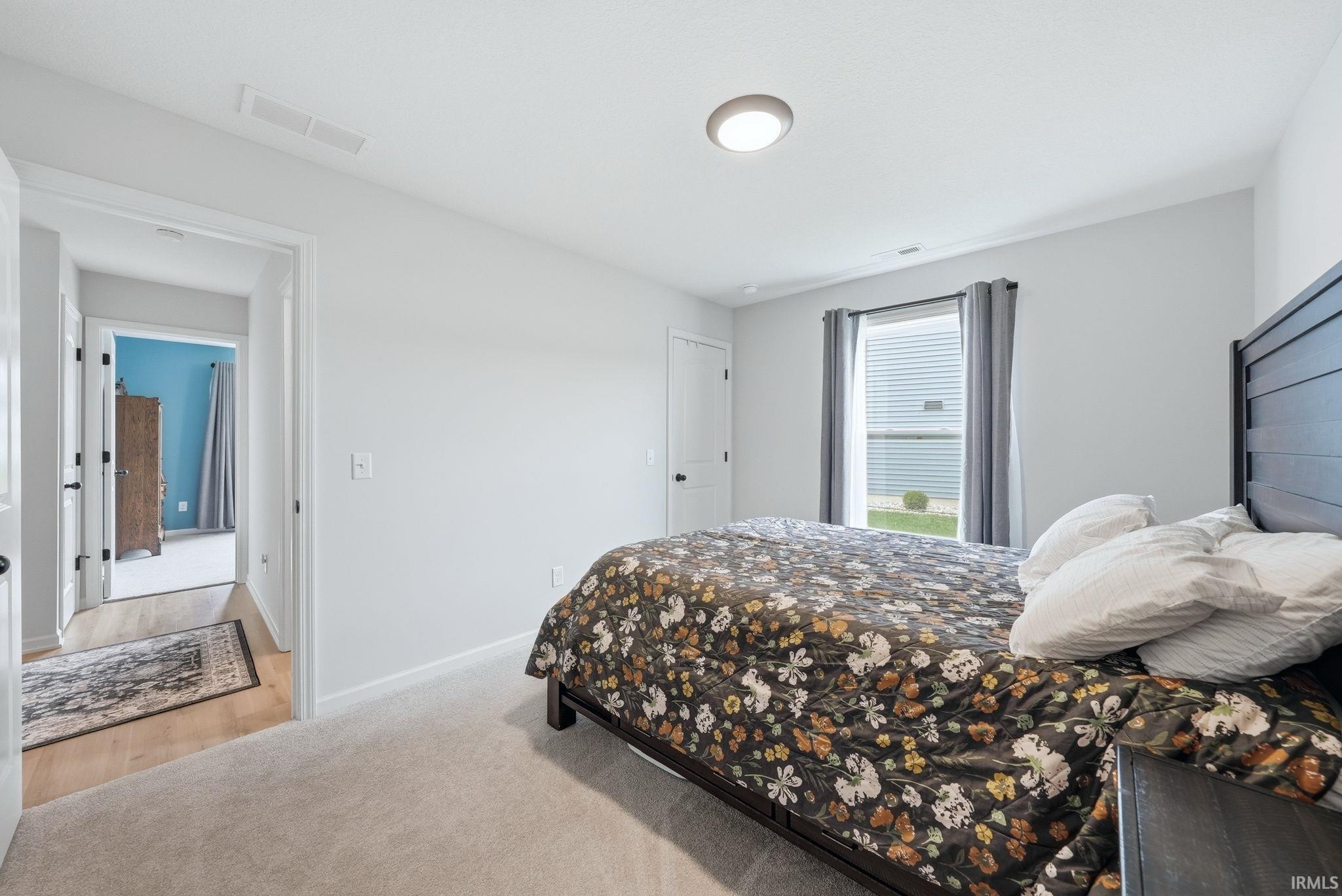 Carpeted room with light gray walls, featuring a window with blinds and gray curtains, a flush-mount ceiling light, and a doorway opening to a hall with wood-finish flooring