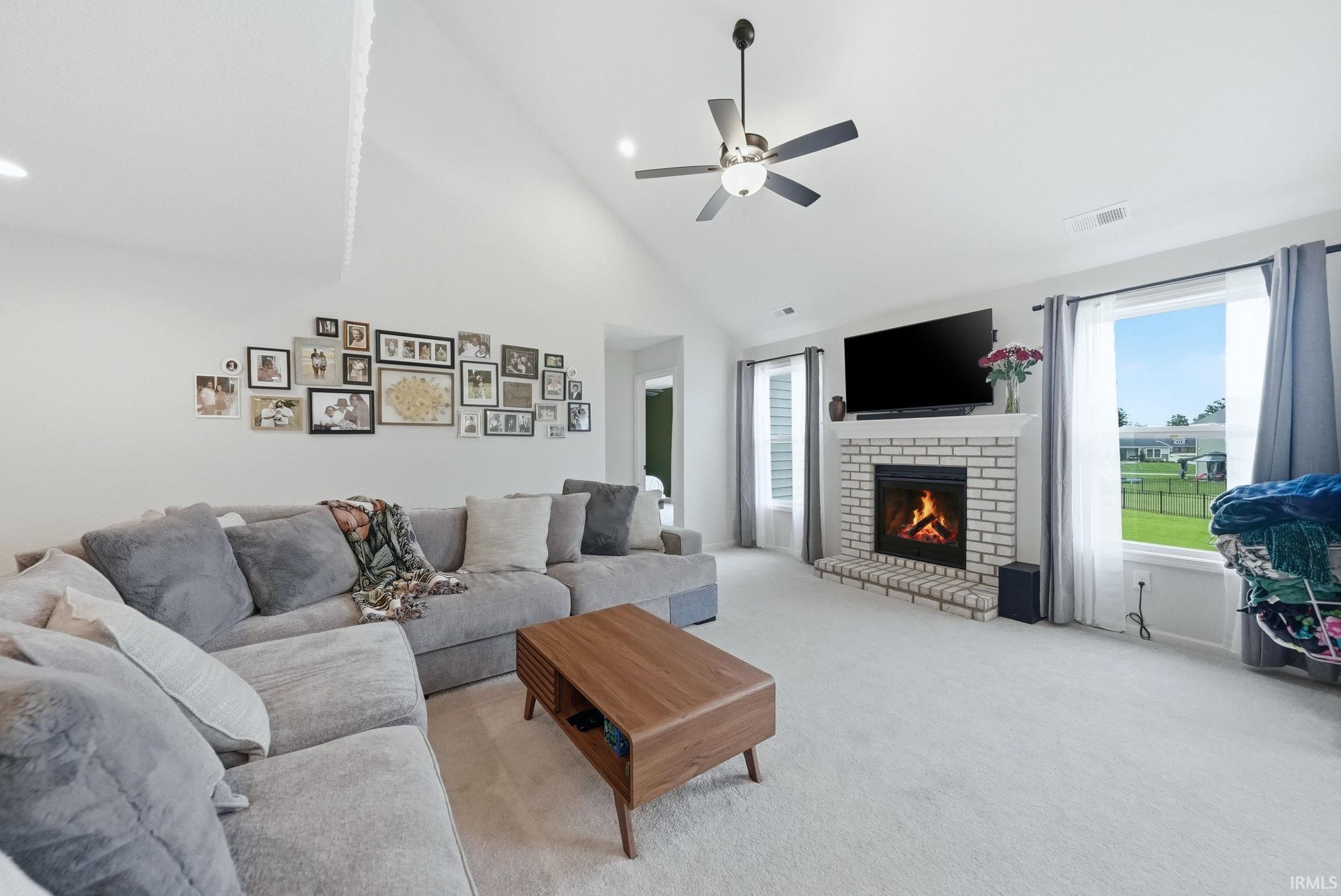 Vaulted ceiling living area featuring a white brick fireplace with a mantle, light-colored carpeting, a ceiling fan with integrated lighting, and multiple windows