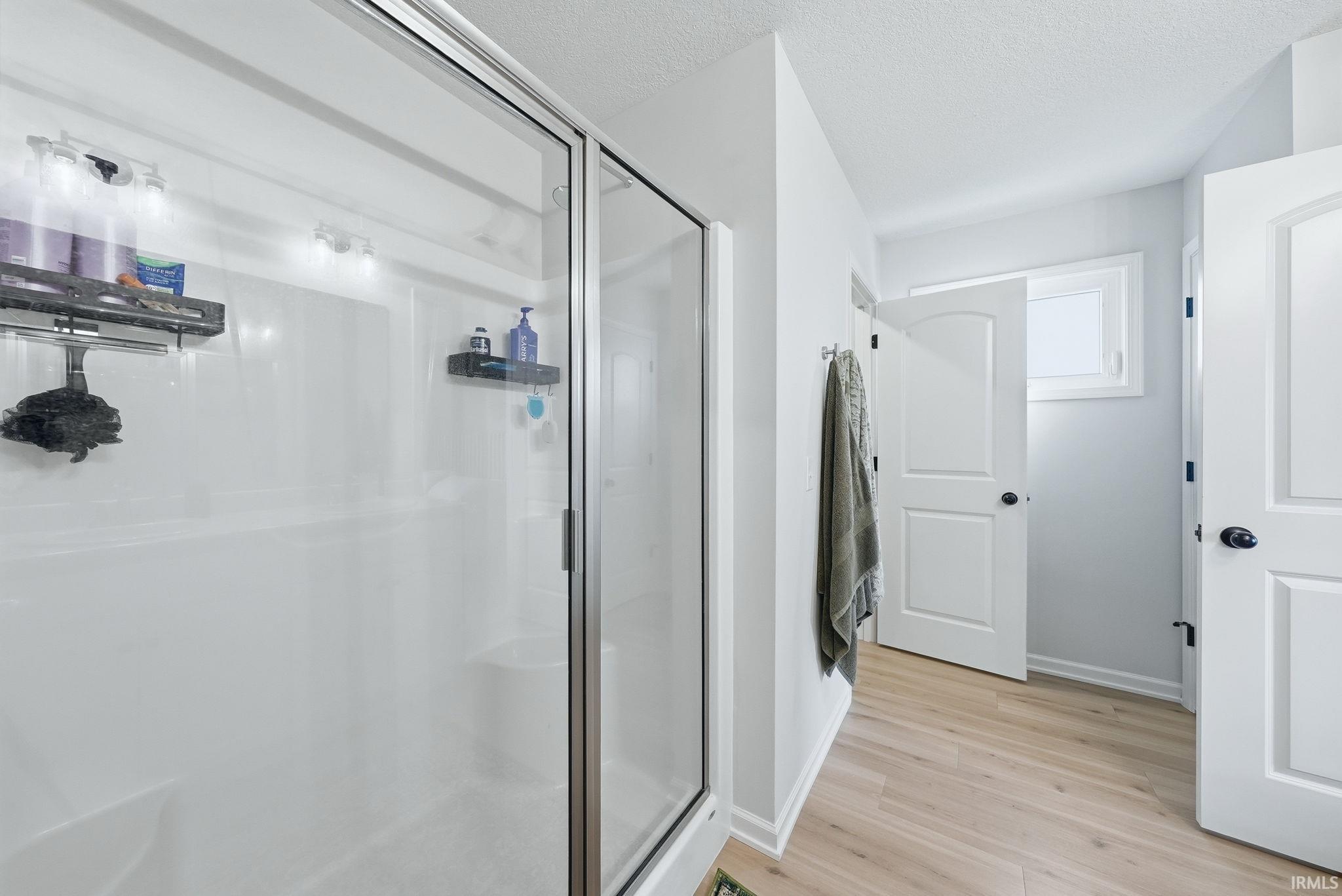 Walk-in shower with clear glass enclosure and integrated shelving, light wood-finish flooring, white doors with black hardware, and light-colored walls