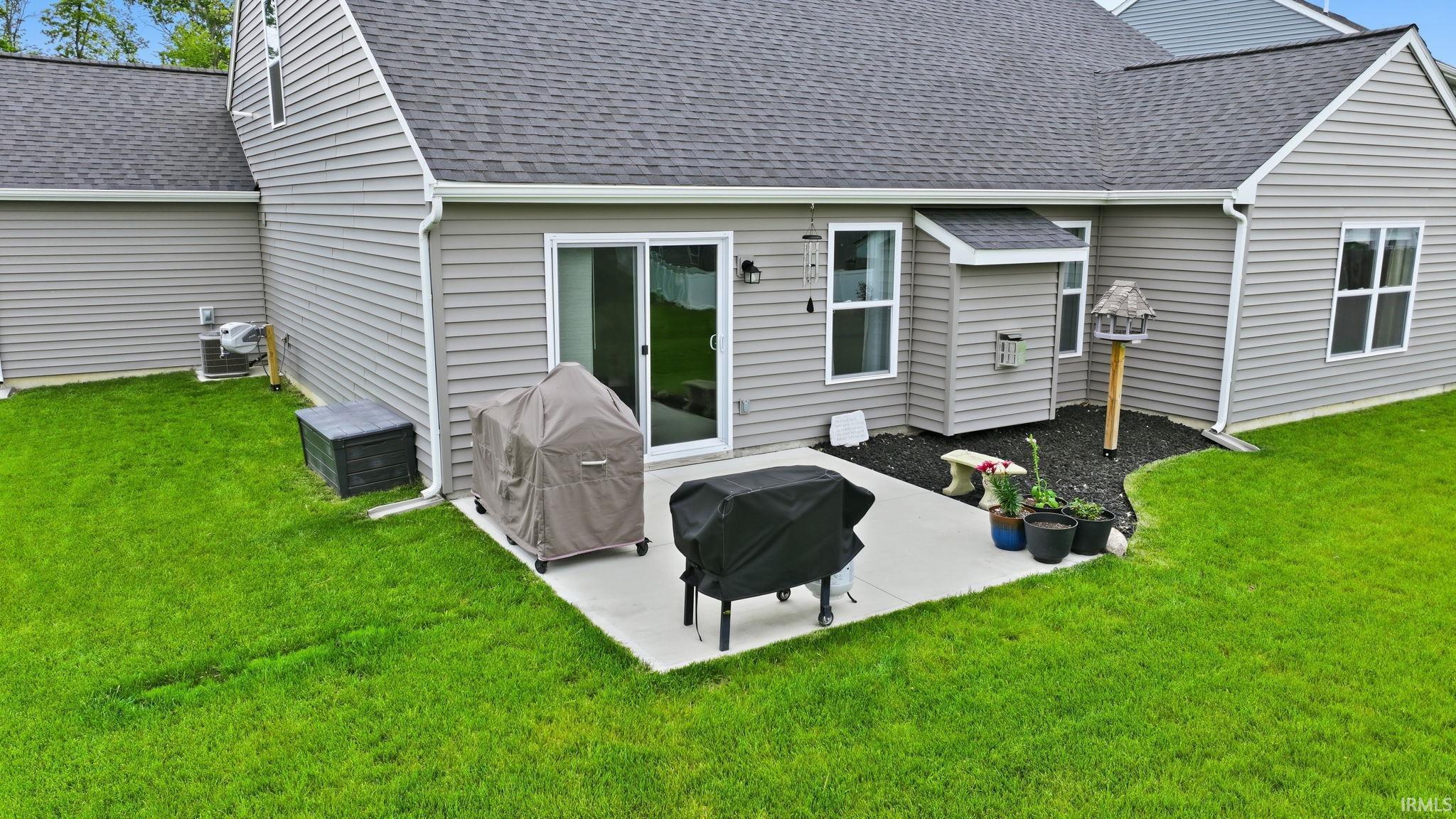 Expansive rear exterior featuring a concrete patio, sliding glass door entry, grey siding, dark shingle roof, and lush green lawn