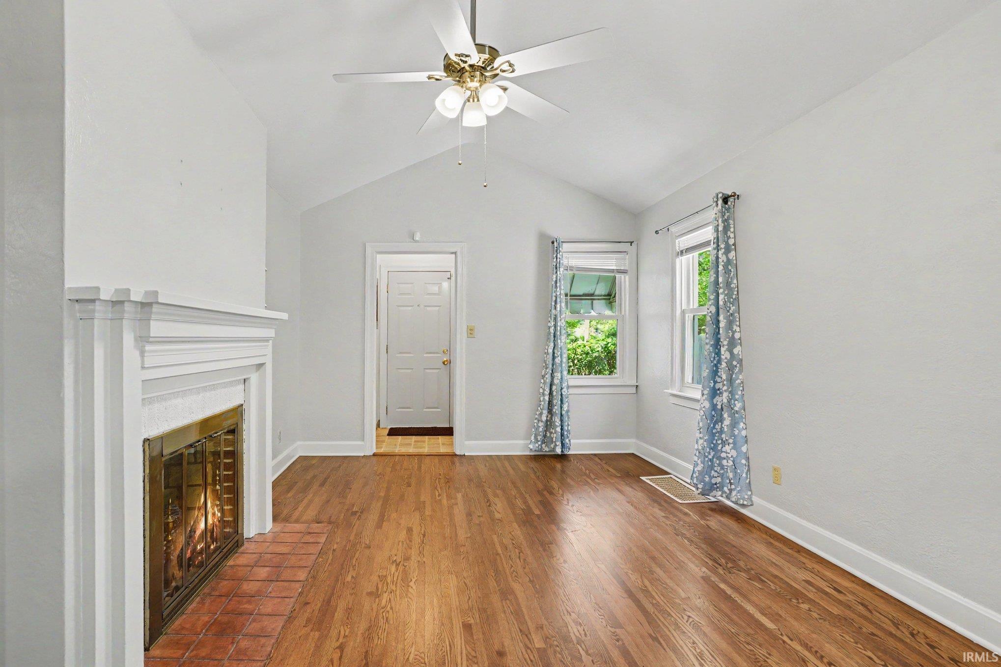 Living room featuring a fireplace with a white mantel and brick hearth, wood-finish flooring, vaulted ceiling, and a ceiling fan with integrated lighting