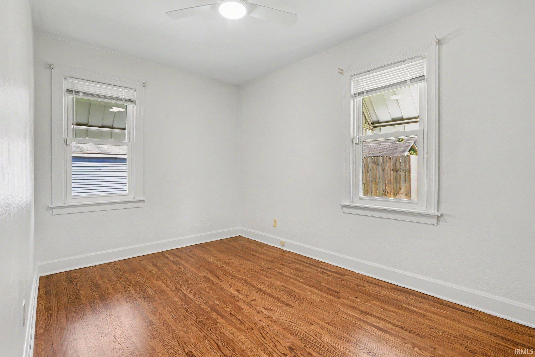 Room featuring wood-finish flooring, two windows with blinds, a ceiling fan, white baseboards, and white walls