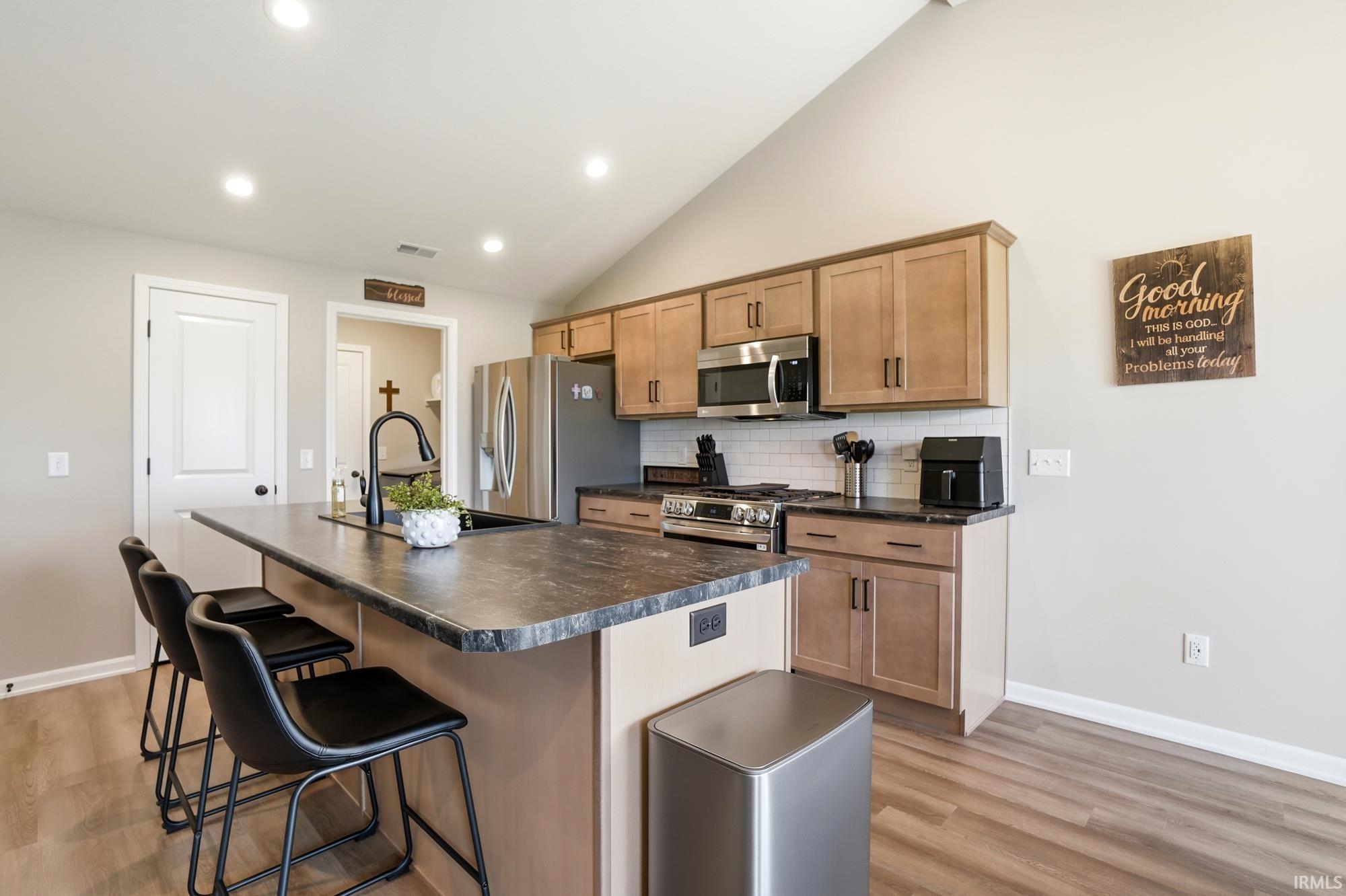 Kitchen featuring a vaulted ceiling with recessed lighting, an island with seating, a black gooseneck faucet, light wood-finish cabinetry, and stainless steel appliances