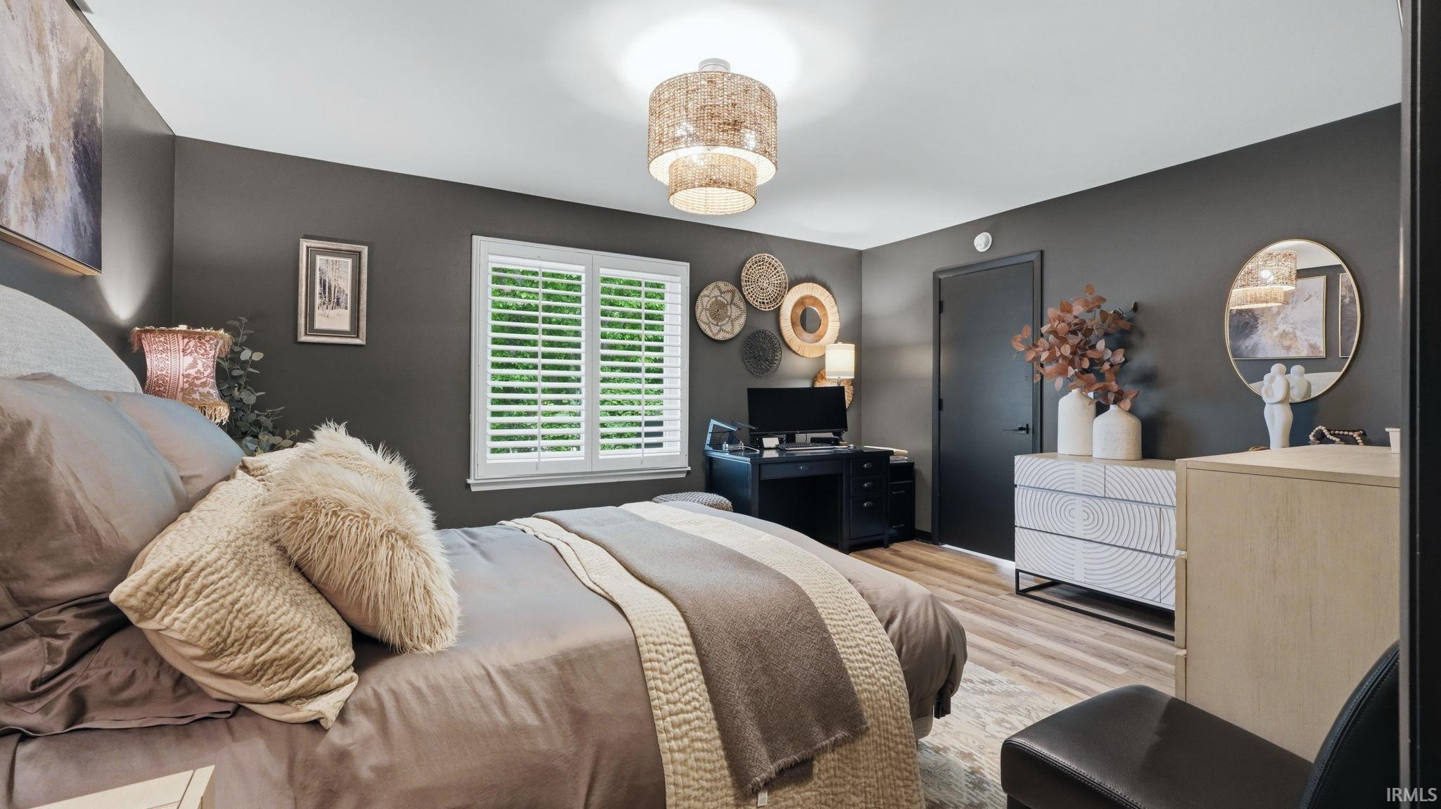 Spacious bedroom featuring wood-finish flooring, a bright window with white plantation shutters, and a modern ceiling light fixture