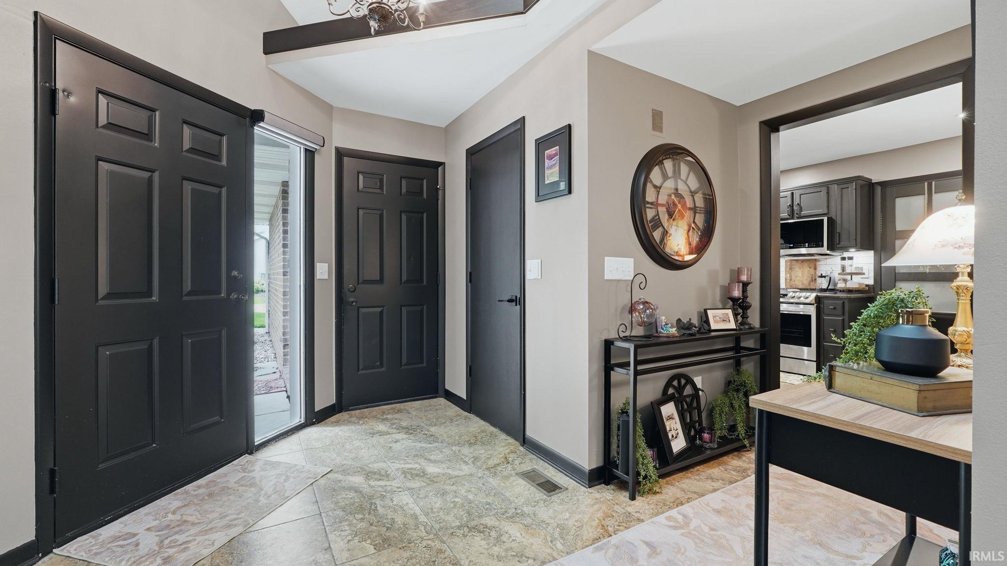 Entryway featuring dark paneled doors, light-toned flooring, and a clear sightline into the kitchen with stainless steel appliances