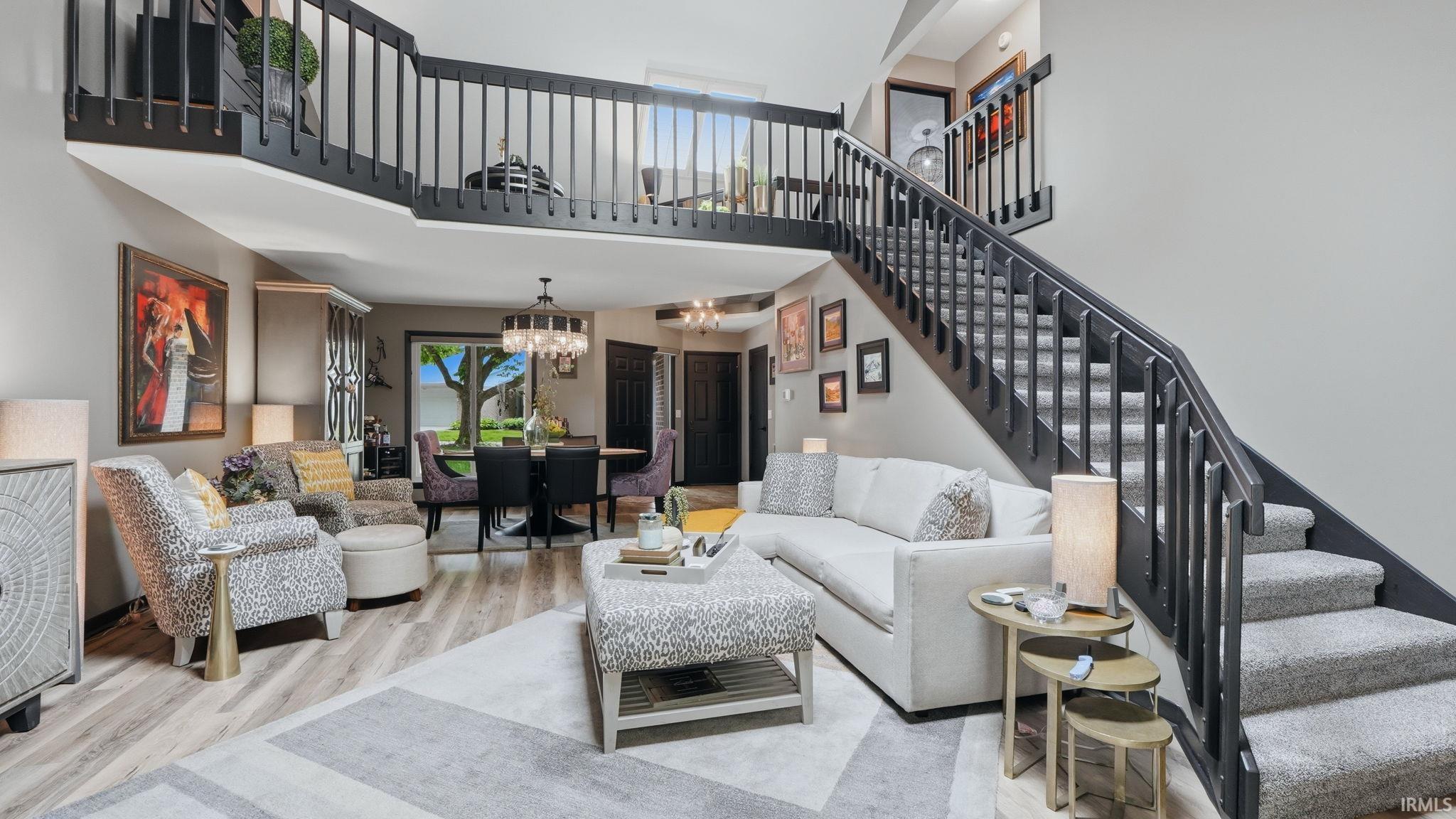 Open-concept living area with wood-finish flooring, a carpeted staircase with dark railings, a dining area with a chandelier, and a balcony overlooking the main space