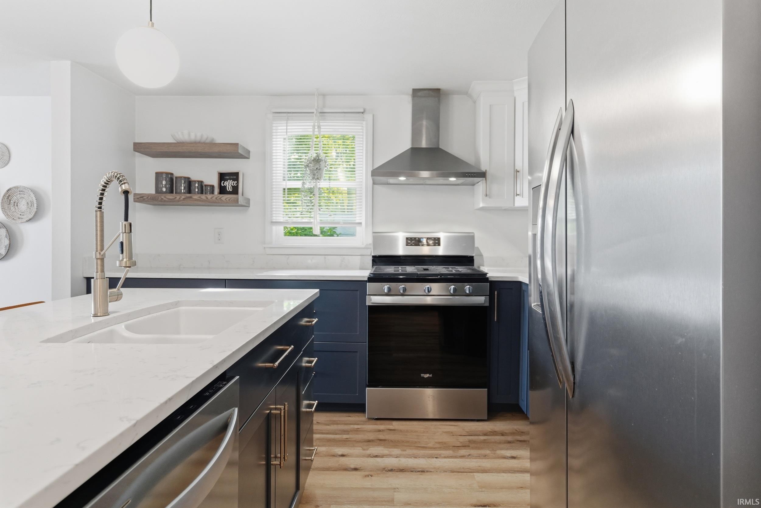 Kitchen featuring a large island with an undermount sink and a pull-down faucet, quartz countertops, dark blue cabinetry, wood-finish flooring, and stainless steel appliances including a range with an overhead hood