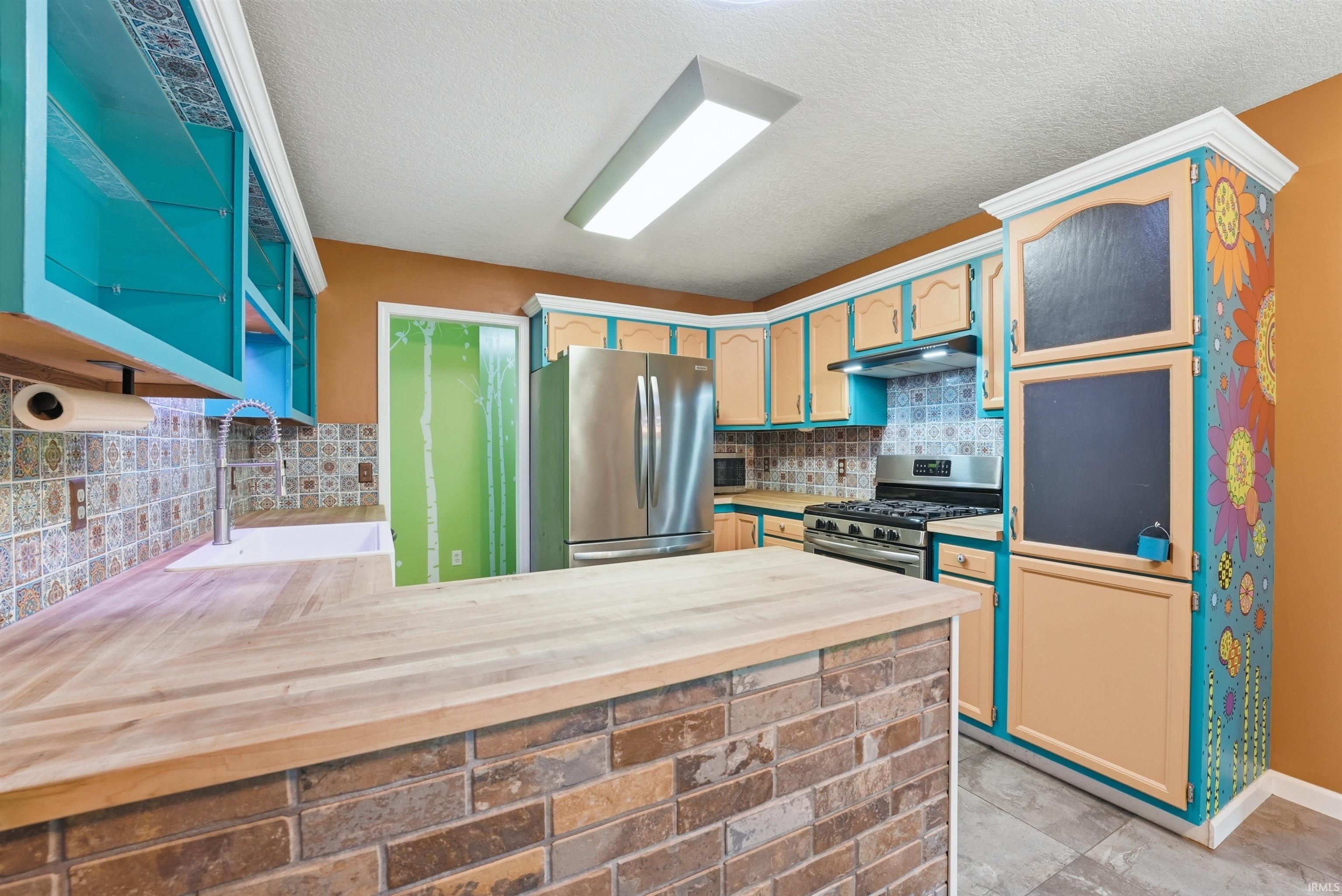 Kitchen featuring a brick-faced island with wood-finish countertop, stainless steel appliances, and patterned tile backsplash