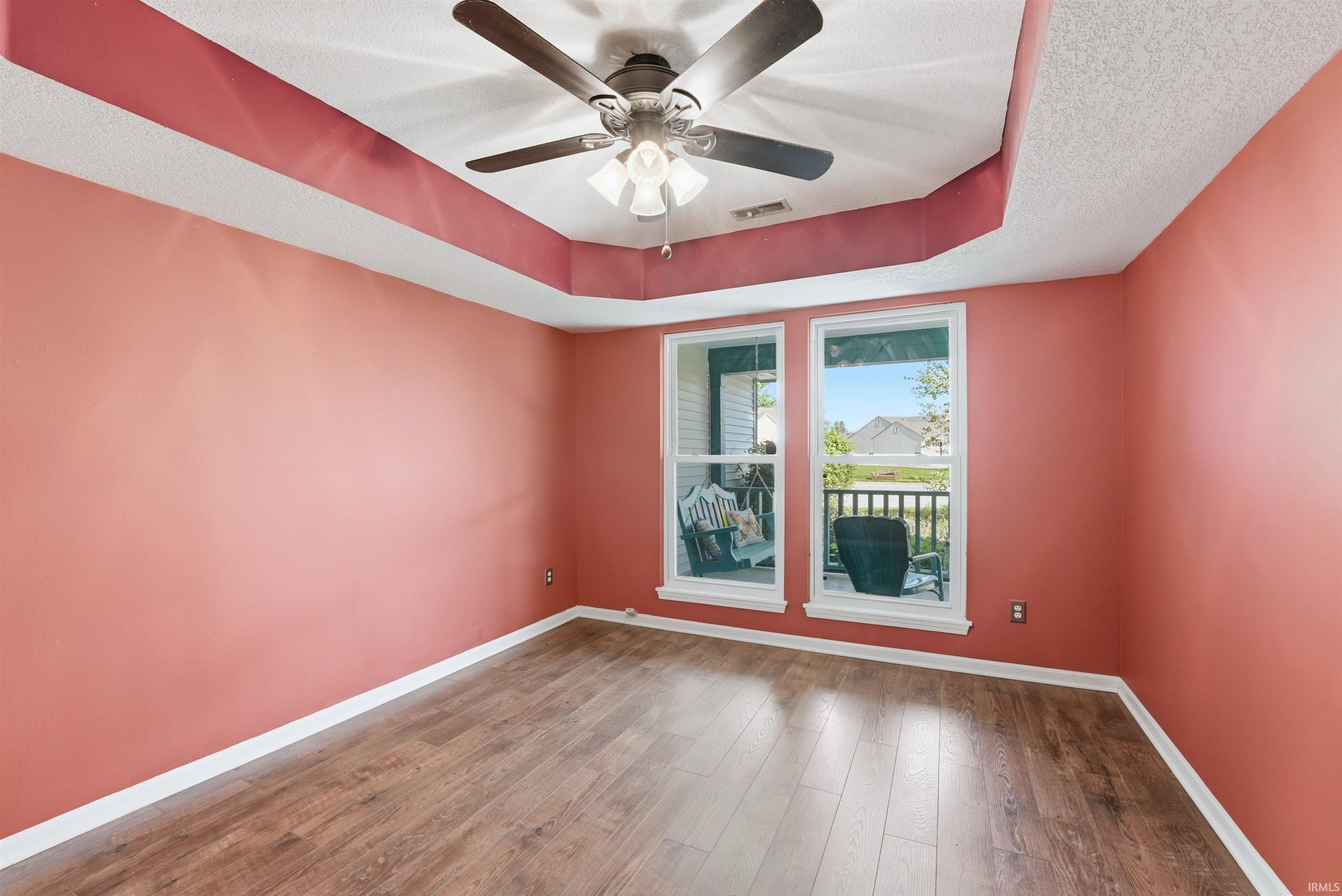 Spacious room featuring wood-finish flooring, a tray ceiling with a ceiling fan, and two large windows