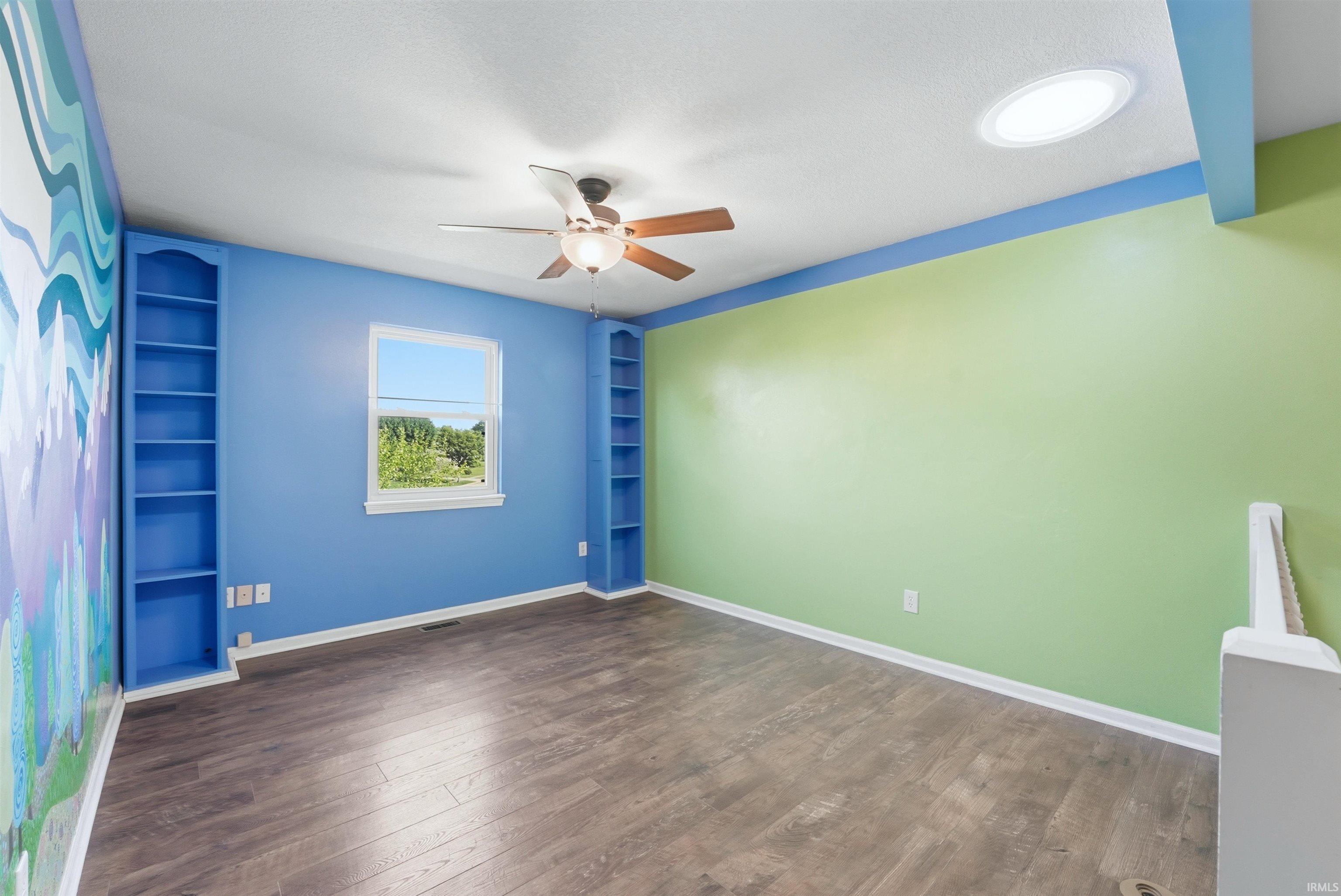 Room featuring wood-finish flooring, two built-in shelving units, a single window, and a ceiling fan with integrated lighting