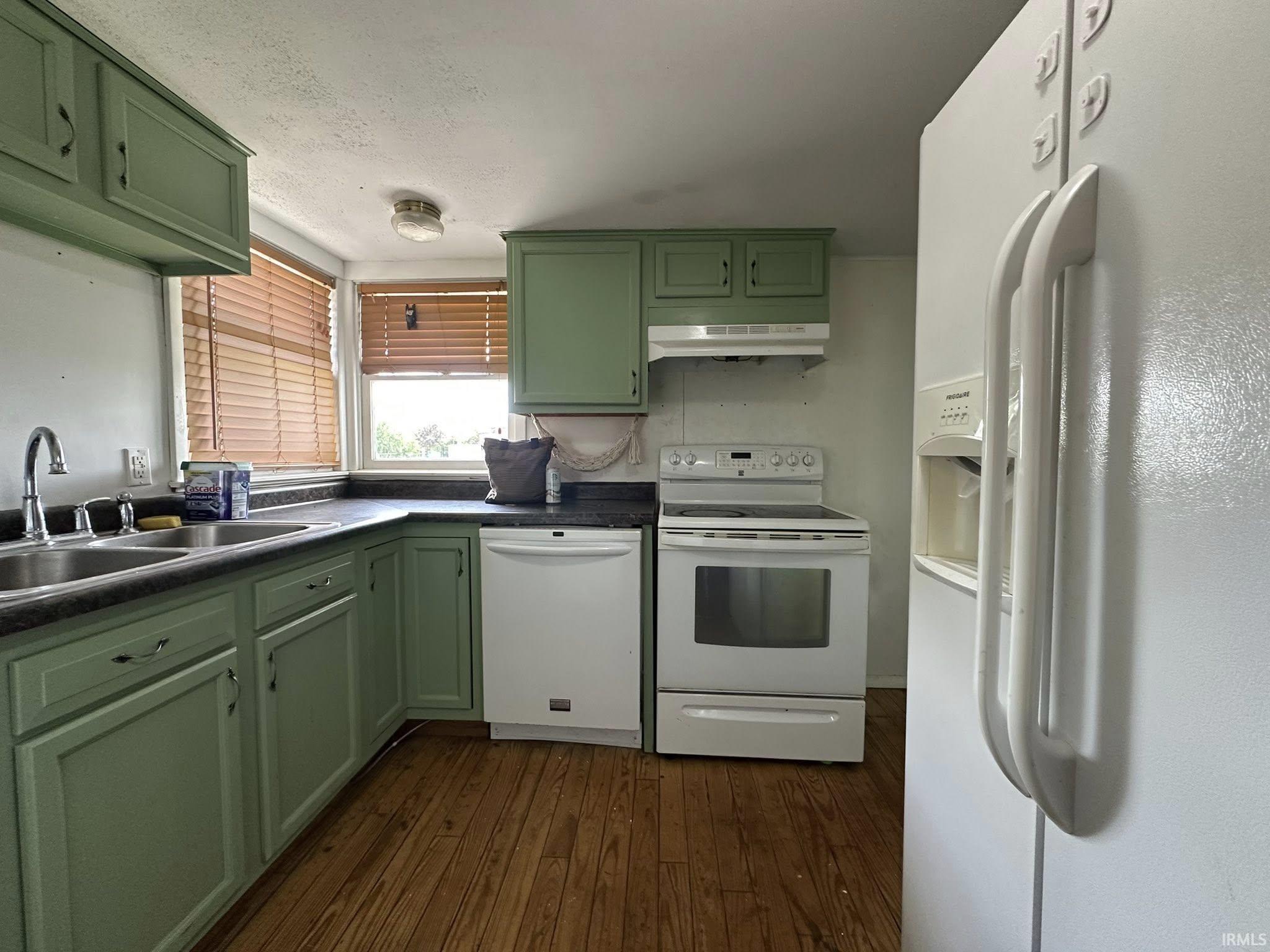 Kitchen featuring wood-finish flooring, green cabinetry, a double basin stainless steel sink, dark countertops, and white appliances including a refrigerator with an ice and water dispenser, a dishwasher, and a range with an overhead hood