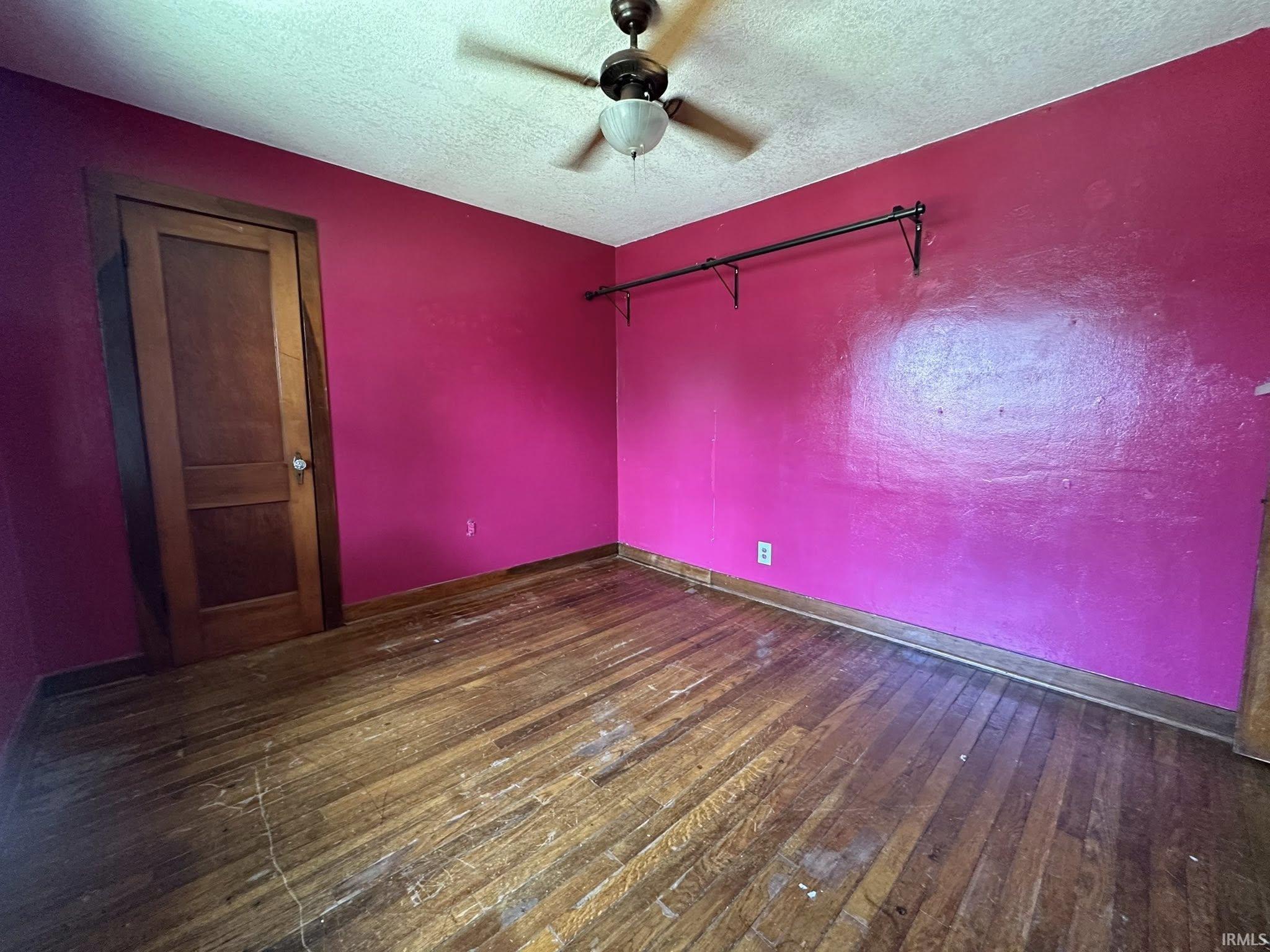 Hardwood flooring, ceiling fan with integrated lighting, wood panel interior door, painted walls, and ceiling-mounted clothing rod