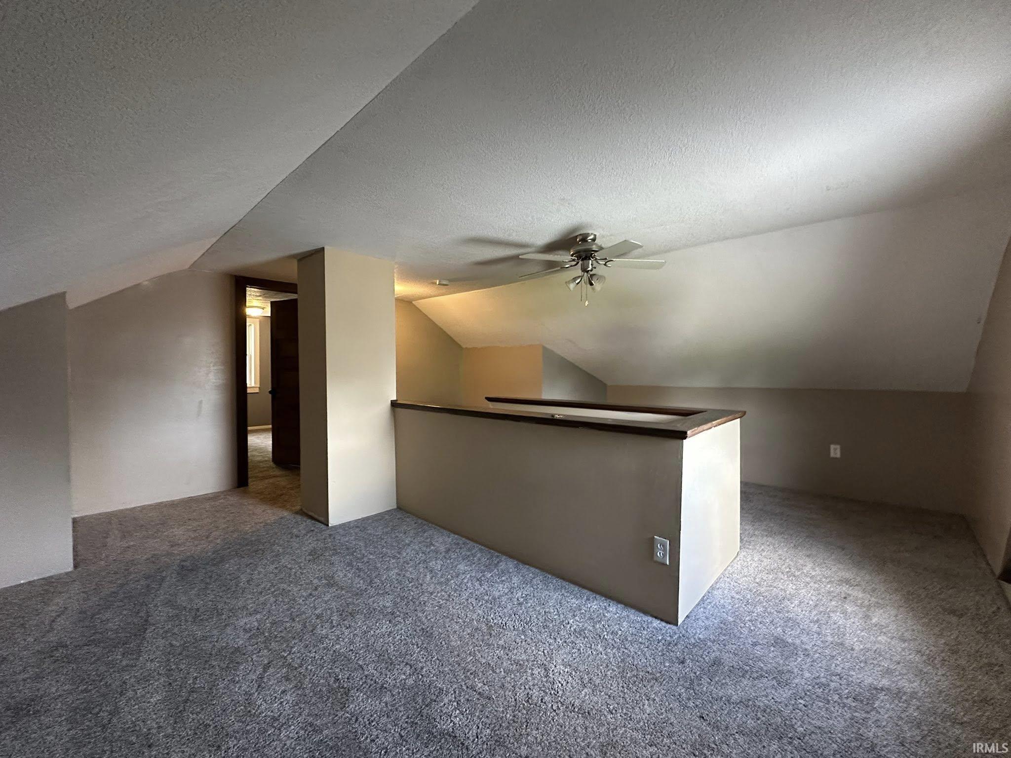 Carpeted bonus room featuring sloped ceilings, a ceiling fan with light fixture, and a partial wall with wood-finish capping