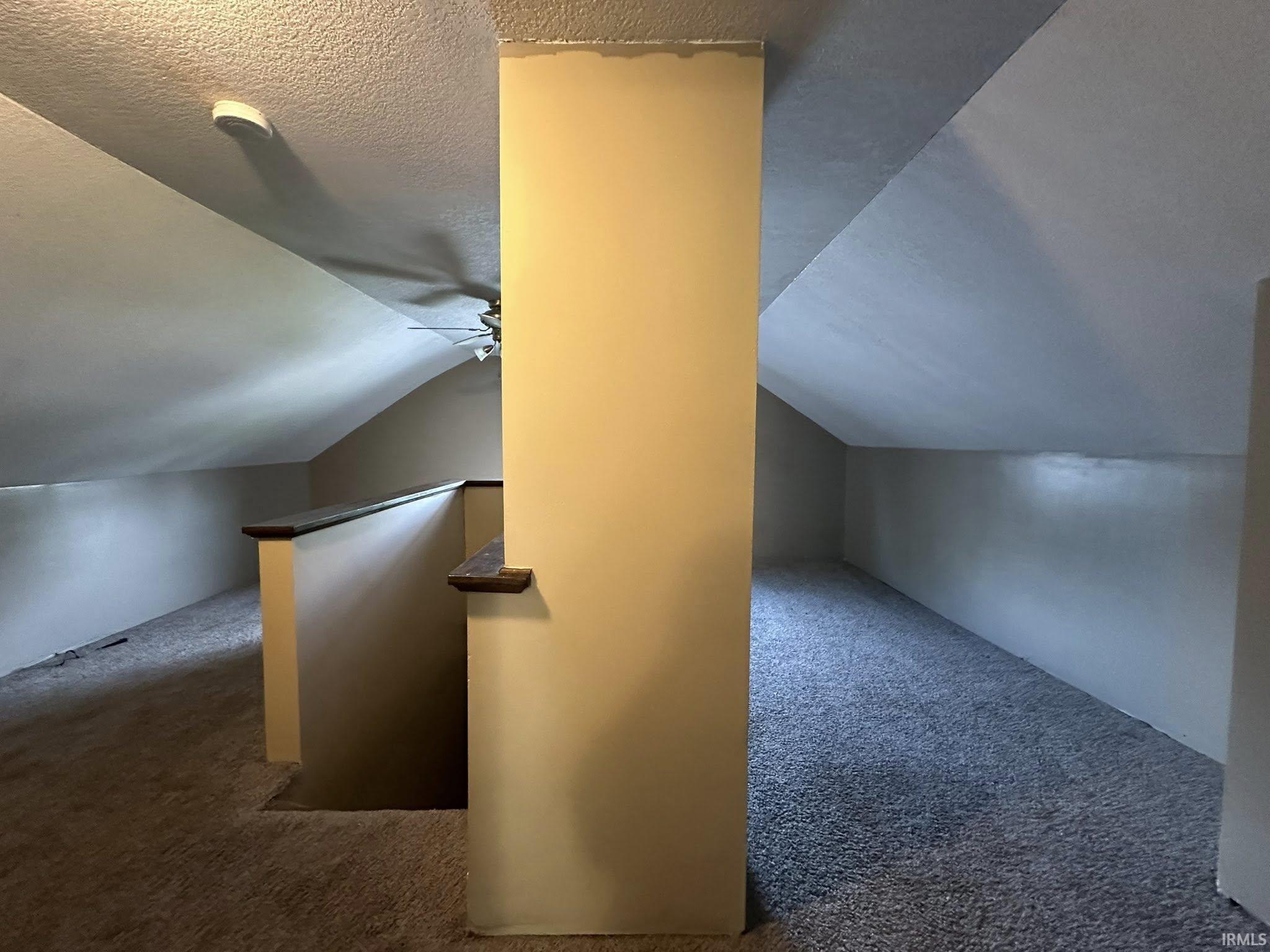 Attic room featuring sloped ceilings, plush carpeting, a partial wall with a dark wood-finish cap, and a ceiling fan