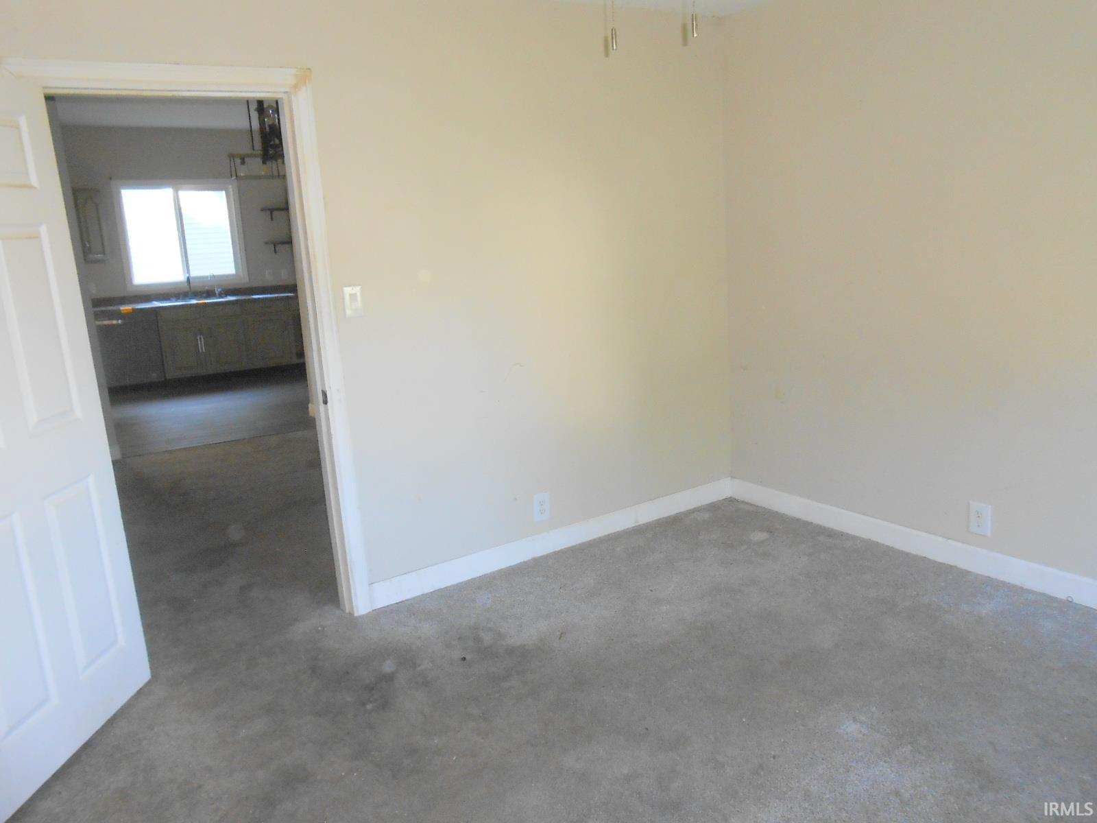 Carpeted room with neutral wall tones and white baseboards, featuring an open doorway revealing a kitchen with a window and lower cabinetry