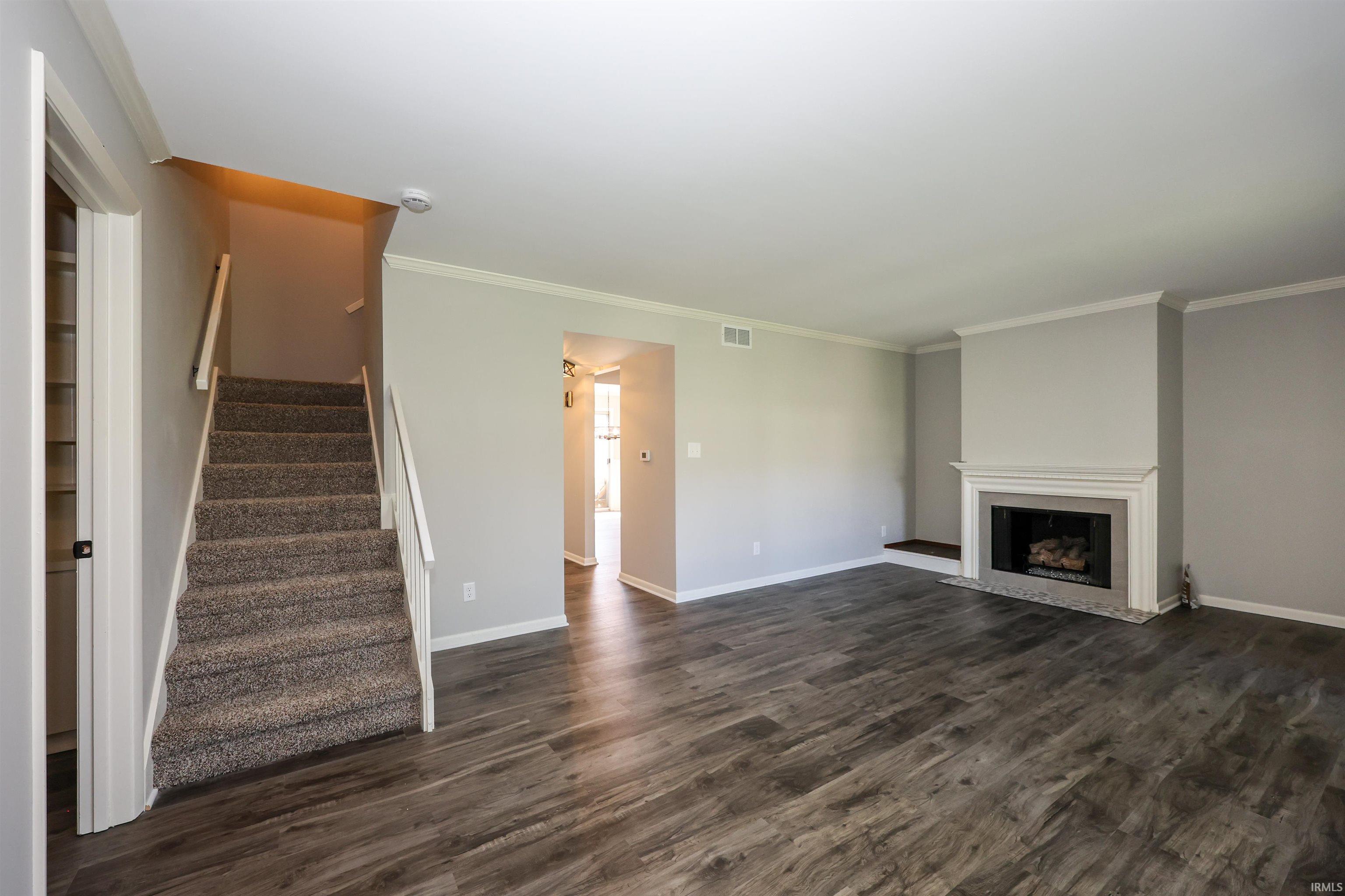 Spacious living area featuring wood-finish flooring, a fireplace with a white mantel, light gray wall paint, white baseboards, and crown molding