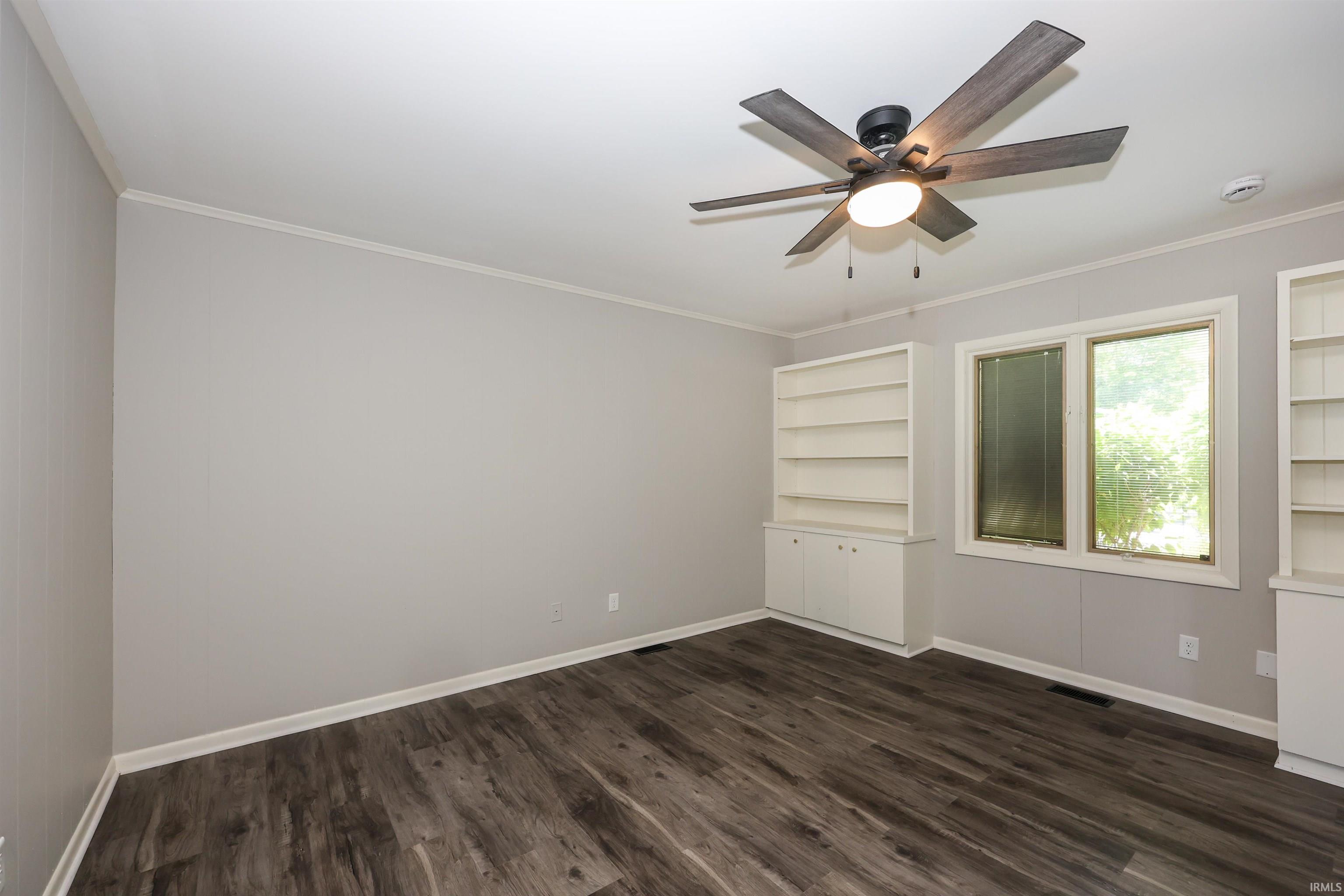 Spacious room featuring wood-finish flooring, two built-in shelving units, a large window, and a ceiling fan with integrated lighting