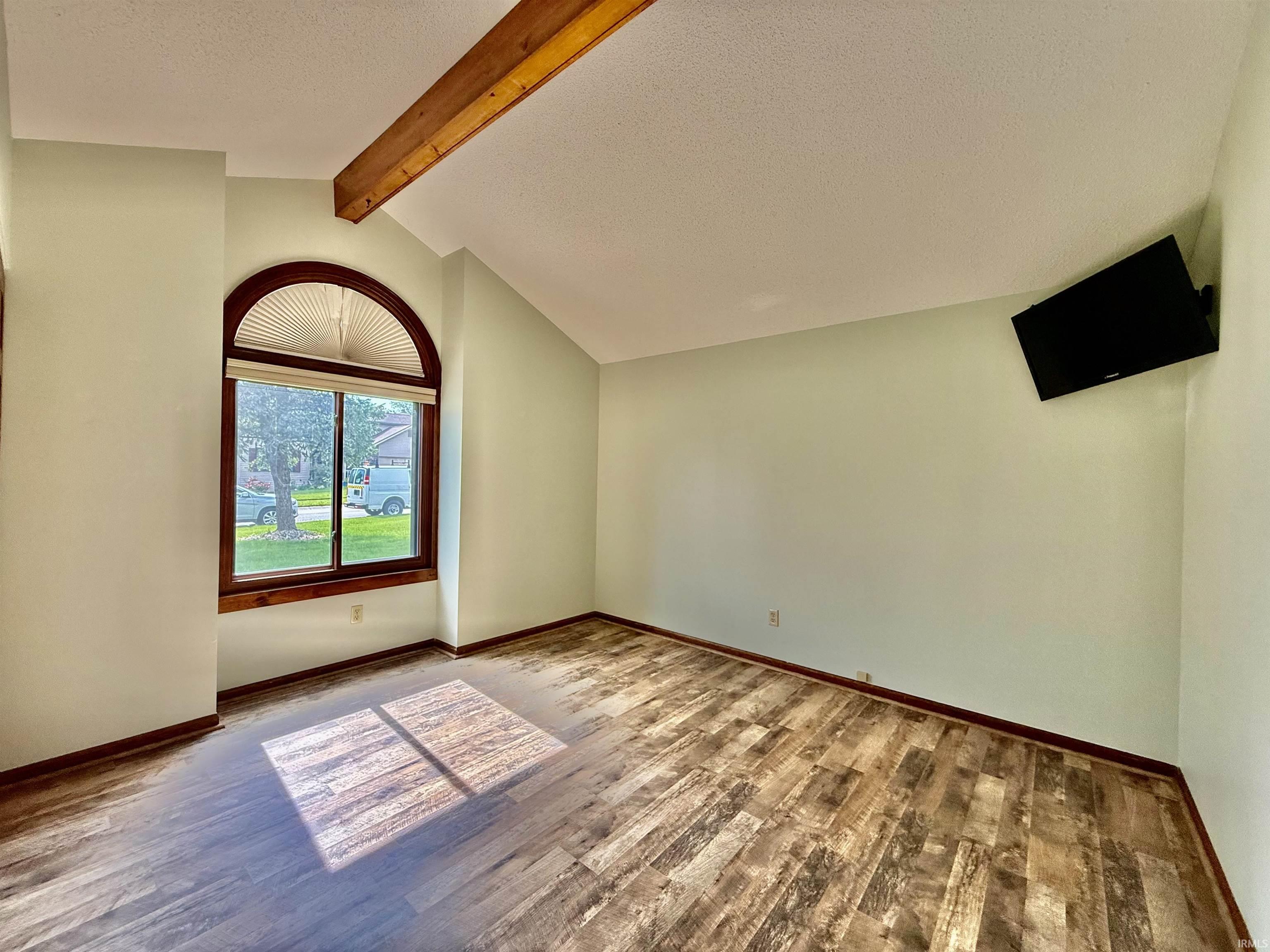Vaulted ceiling with exposed wood beam, arched window with horizontal blinds, wood-finish flooring, and light-toned wall paint