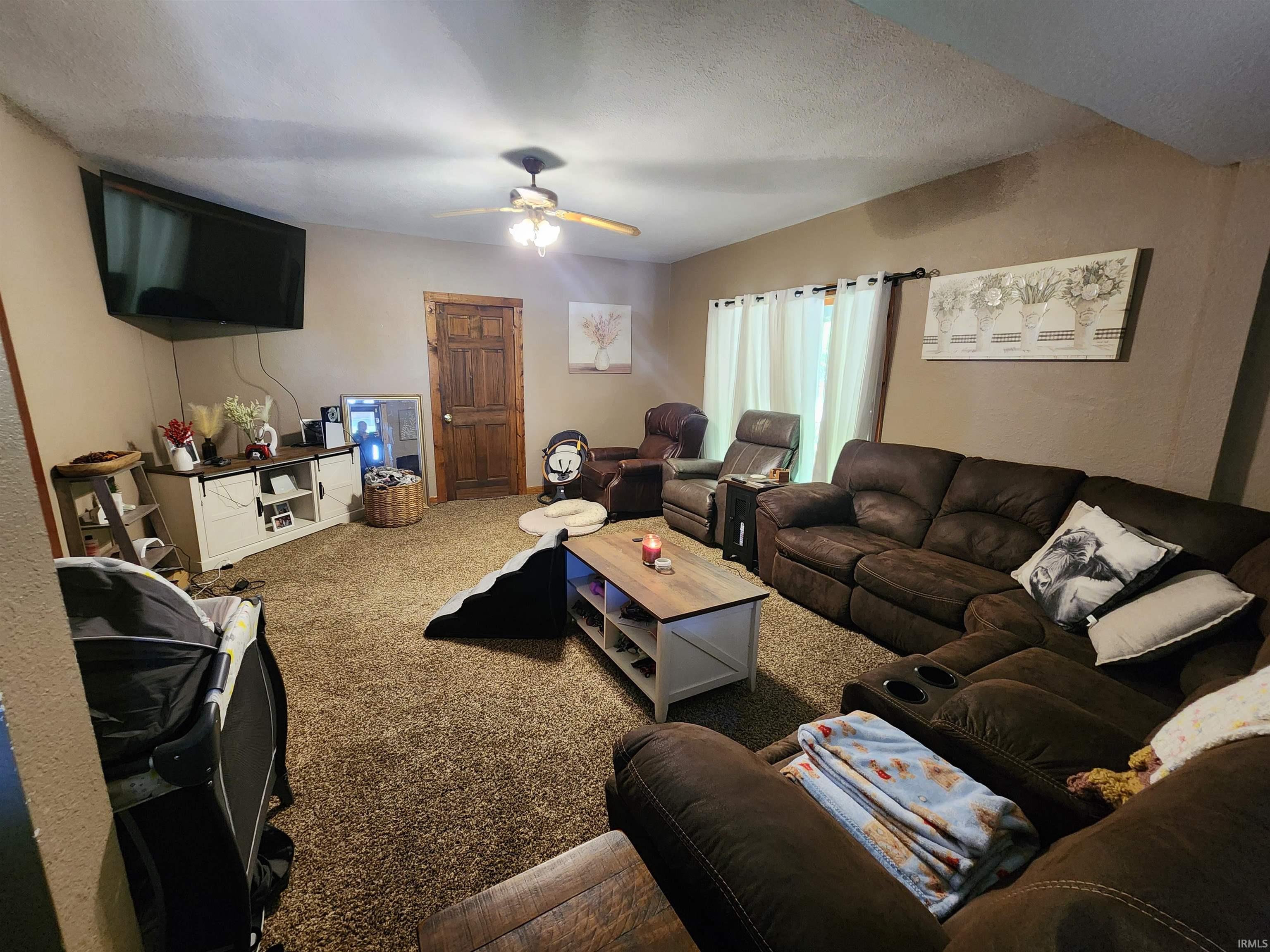 Spacious living area featuring neutral wall tones, textured carpeting, a ceiling fan with light fixture, window with sheer curtains, and a solid wood interior door