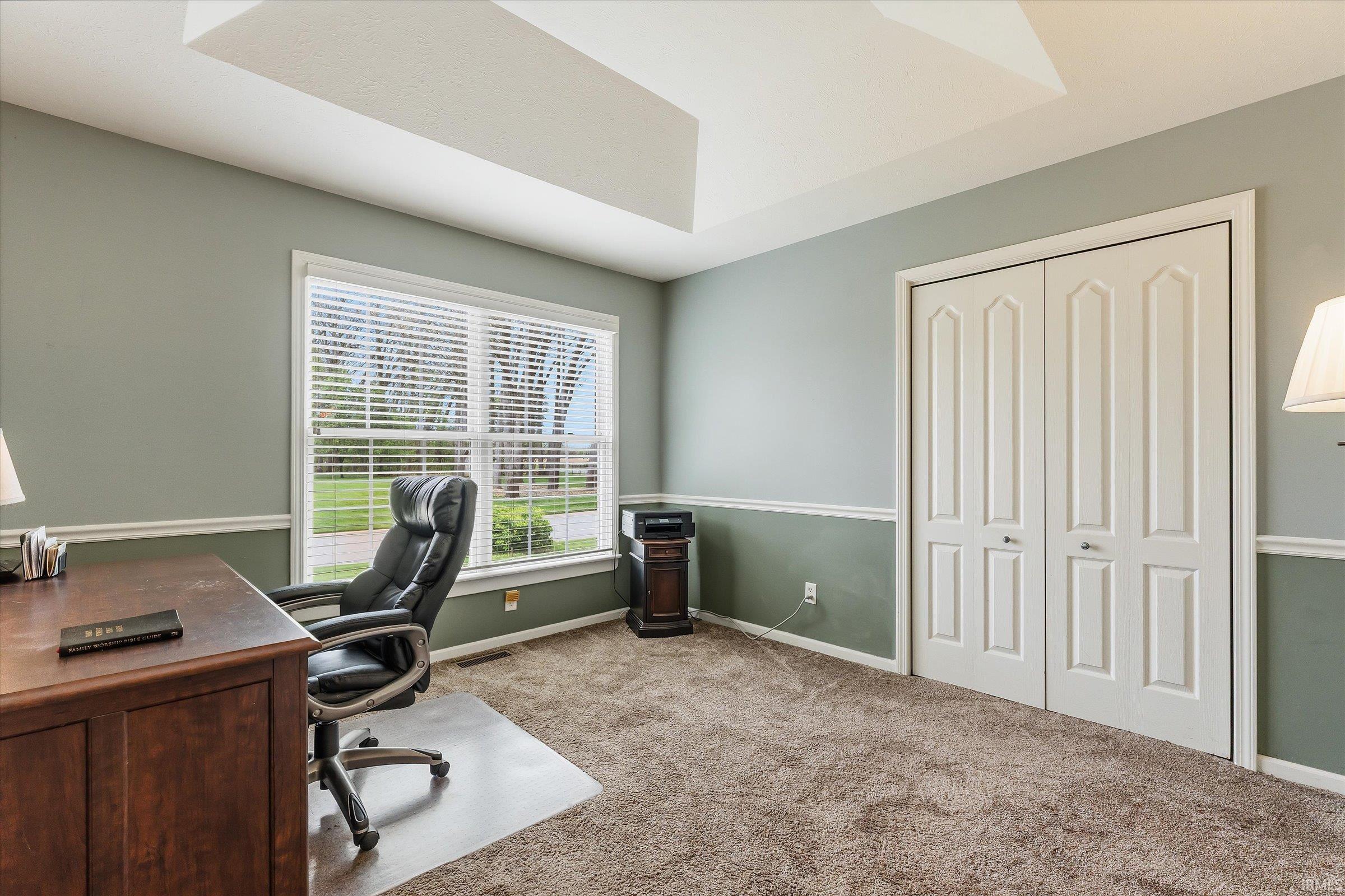 Carpeted room featuring a large window with blinds, tray ceiling, two-tone painted walls with chair rail, and bi-fold closet doors