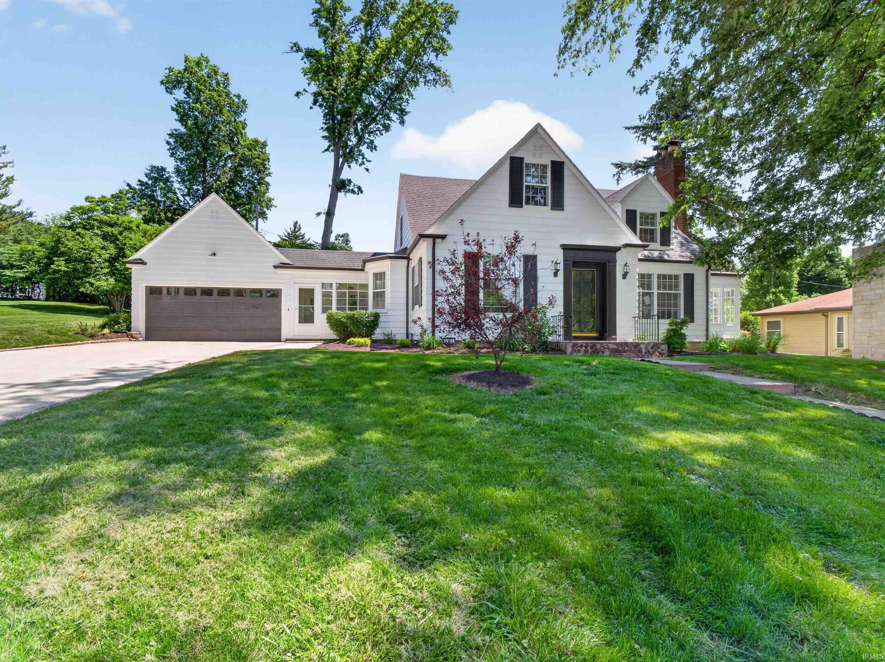 White siding exterior with black shutters, dormer windows, attached garage, stone accents, and mature tree landscaping