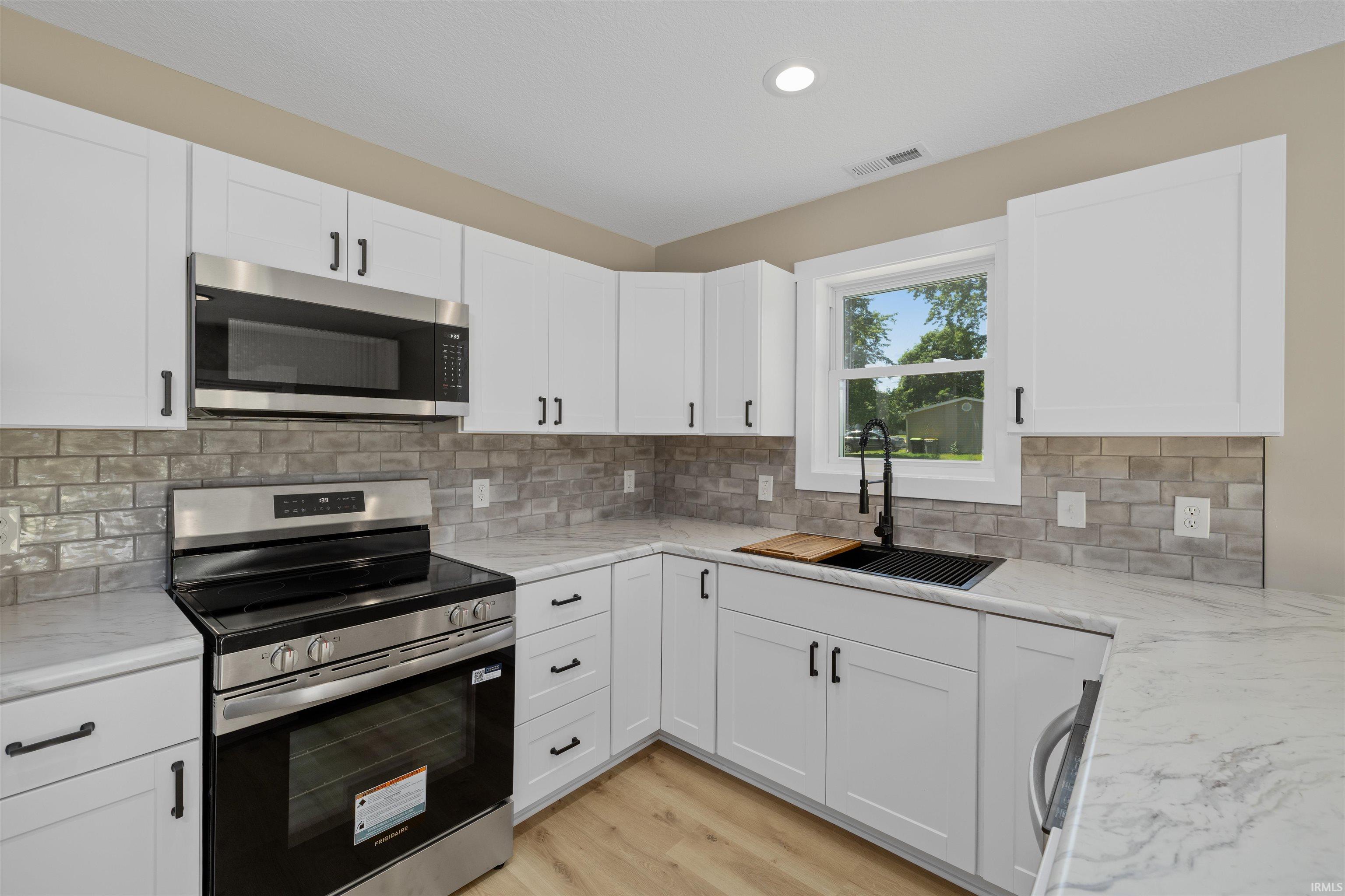 Modern kitchen featuring white shaker-style cabinetry with matte black hardware, stainless steel appliances, light-toned wood-finish flooring, white countertops with gray veining, and a gray brick-patterned backsplash