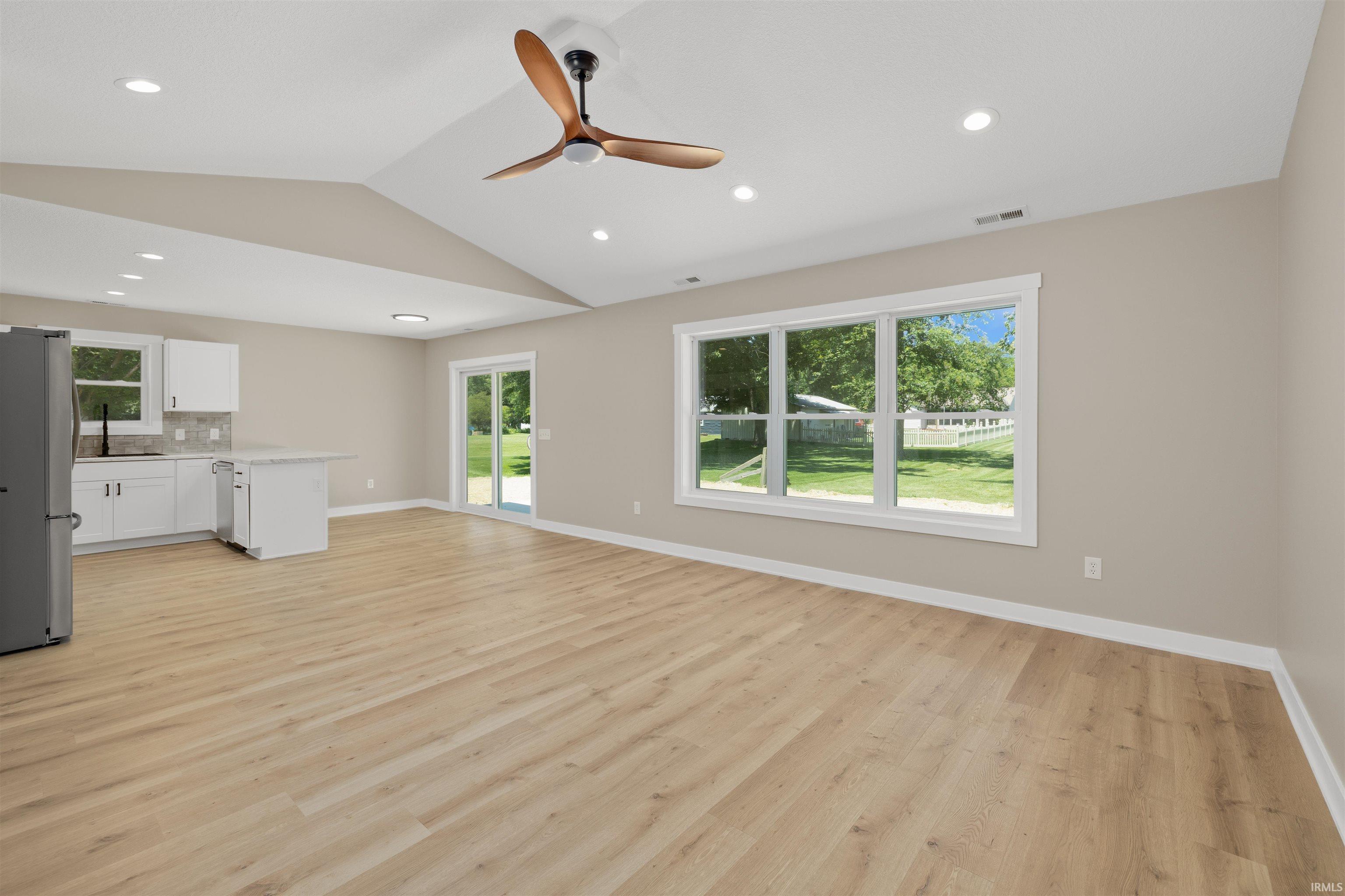 Open-concept living space featuring wood-finish flooring, vaulted ceilings with a ceiling fan, recessed lighting, and a kitchen area with white cabinetry, light countertops, and a tiled backsplash