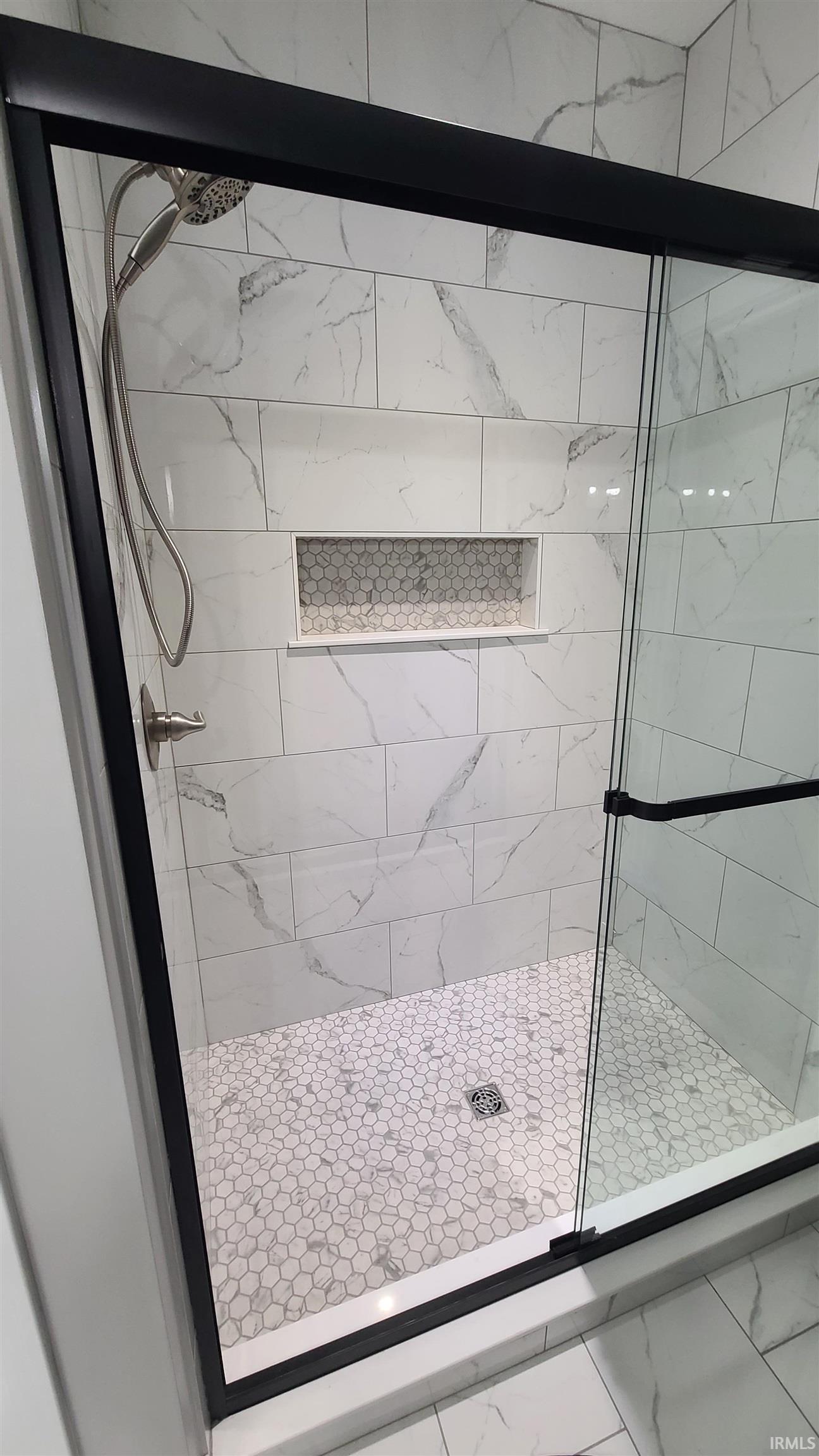 Shower enclosure featuring a black-framed glass door, white rectangular wall tiles with veining, a built-in shower niche with mosaic tile, a handheld shower fixture, and a hexagonal mosaic tile floor