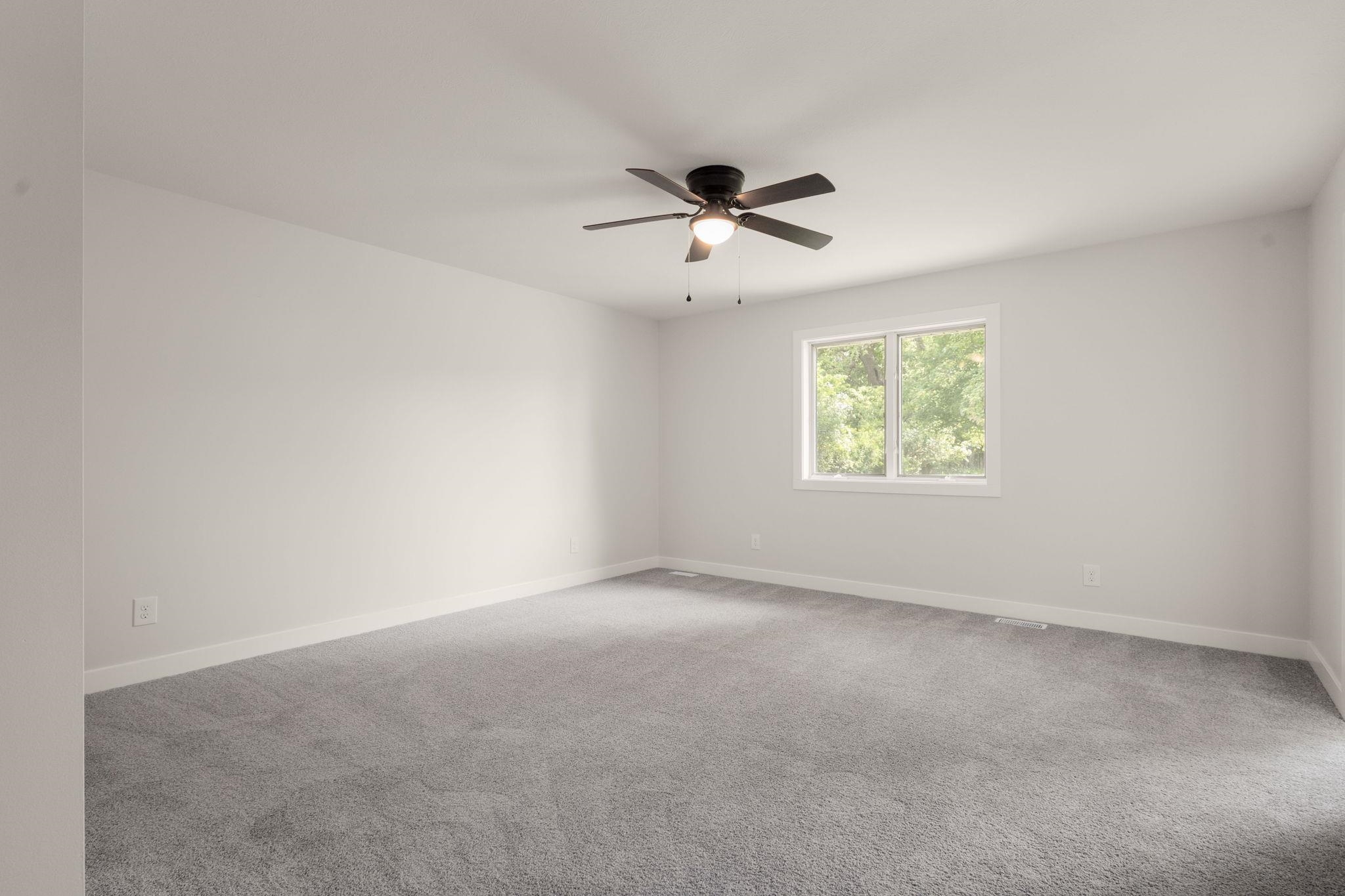 Spacious room featuring light gray carpeting, a multi-pane window with white trim, a ceiling fan with integrated lighting, and white baseboards