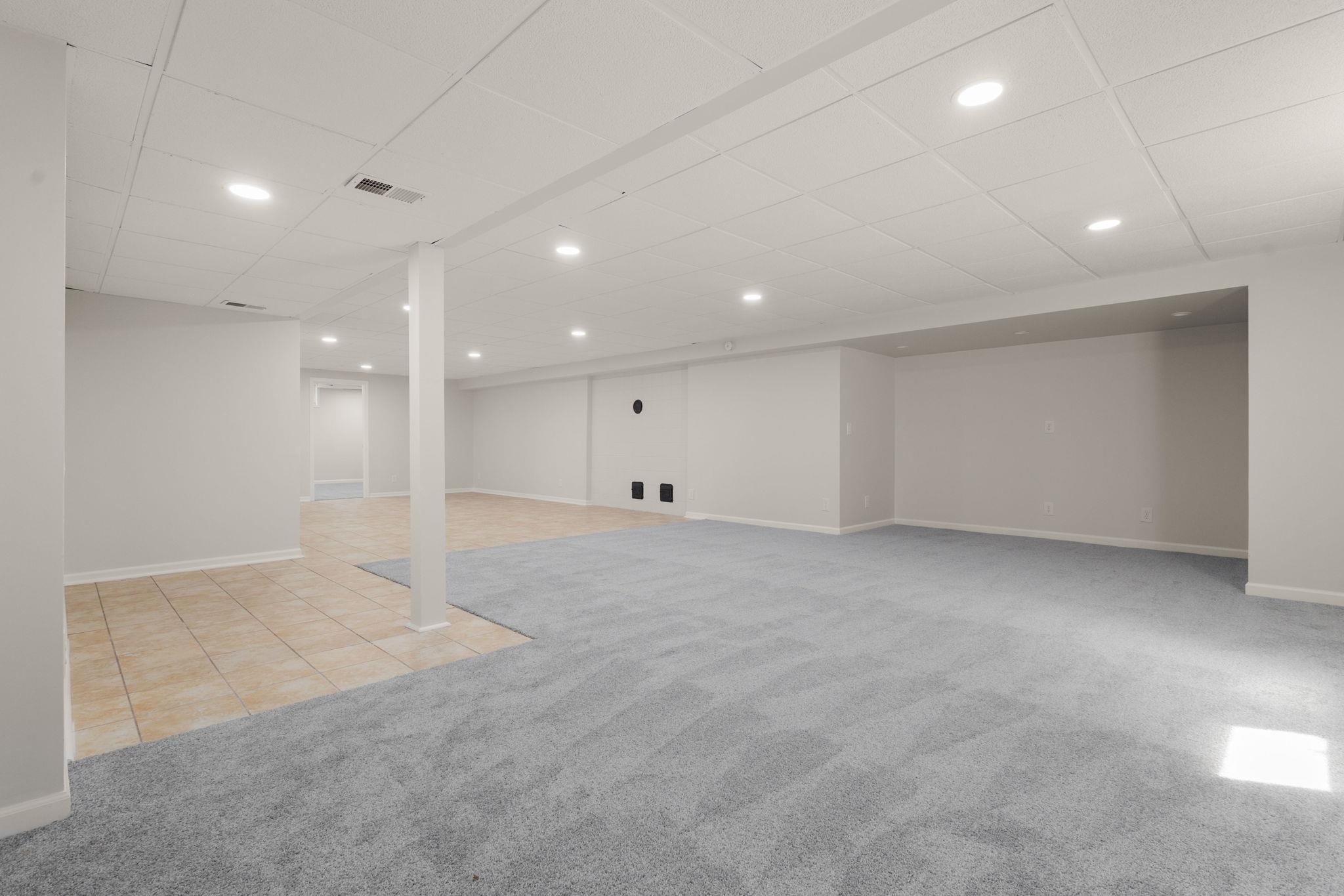 Spacious lower-level area featuring light gray carpeting, neutral wall finishes, a drop ceiling with recessed lighting, and a tiled section with tan square tiles
