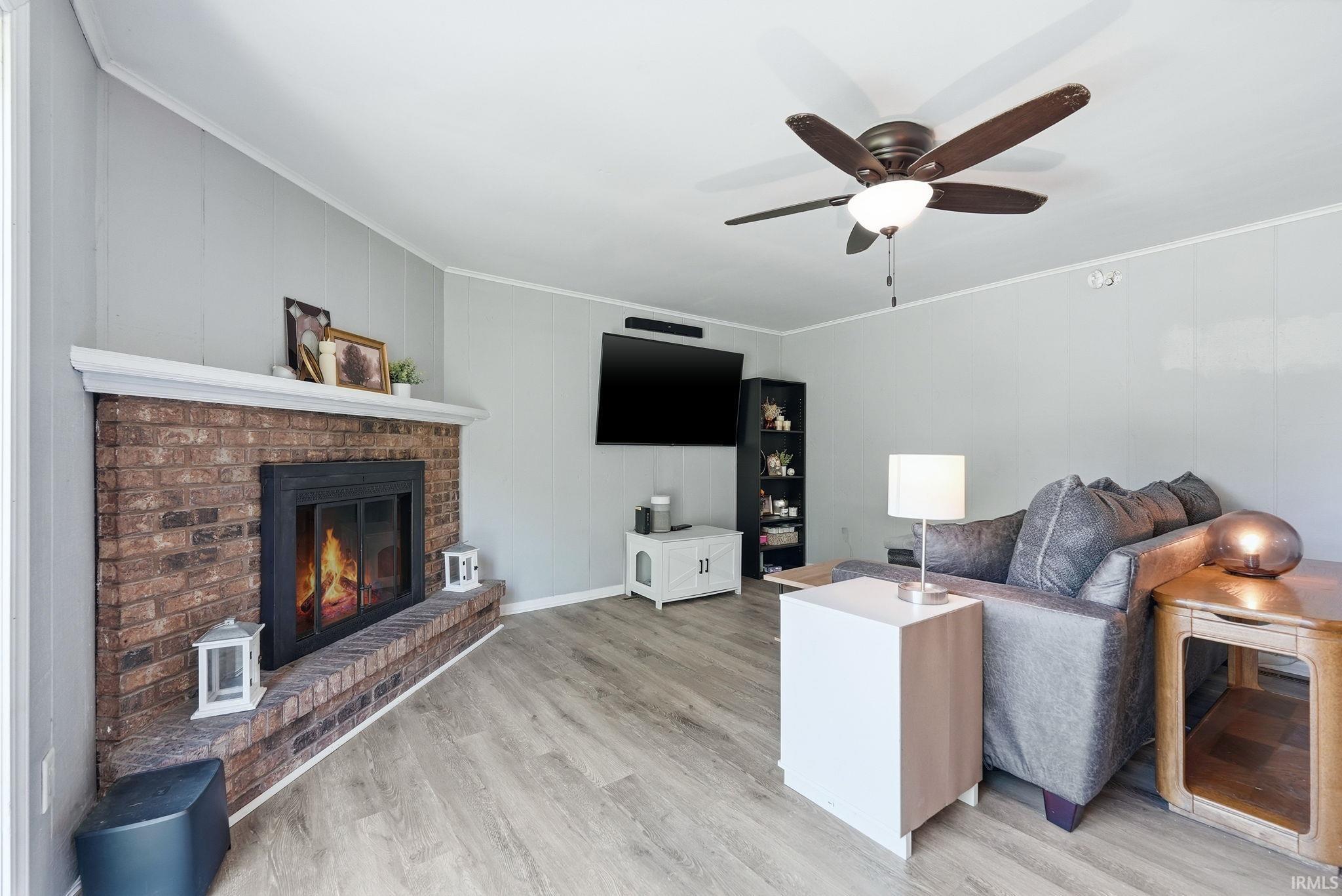 Living area featuring a brick-face fireplace with a white mantle, wood-finish flooring, a ceiling fan, and light gray paneled walls