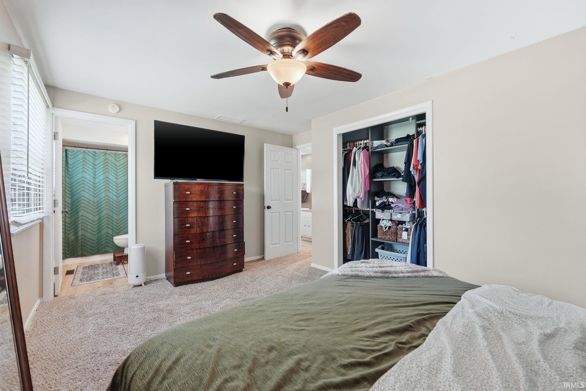 Carpeted bedroom featuring a ceiling fan with light fixture, built-in closet with shelving and hanging rods, and an en-suite bathroom entrance